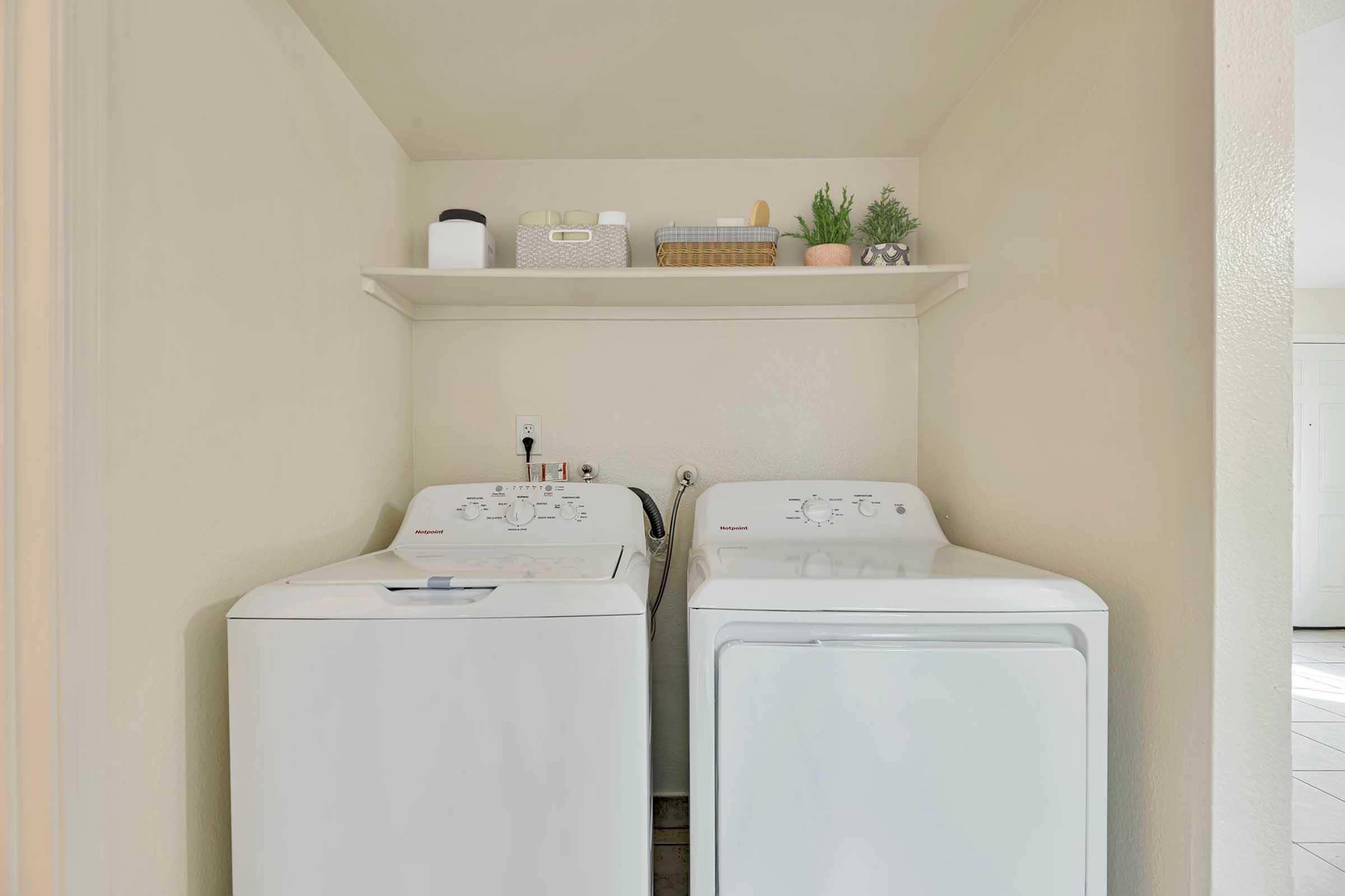 A clean and organized laundry area featuring a top-loading washing machine and a dryer side by side. Above them, a wall-mounted shelf holds neatly arranged laundry supplies and decorative items. The walls are painted in a light color, creating a bright and inviting space.