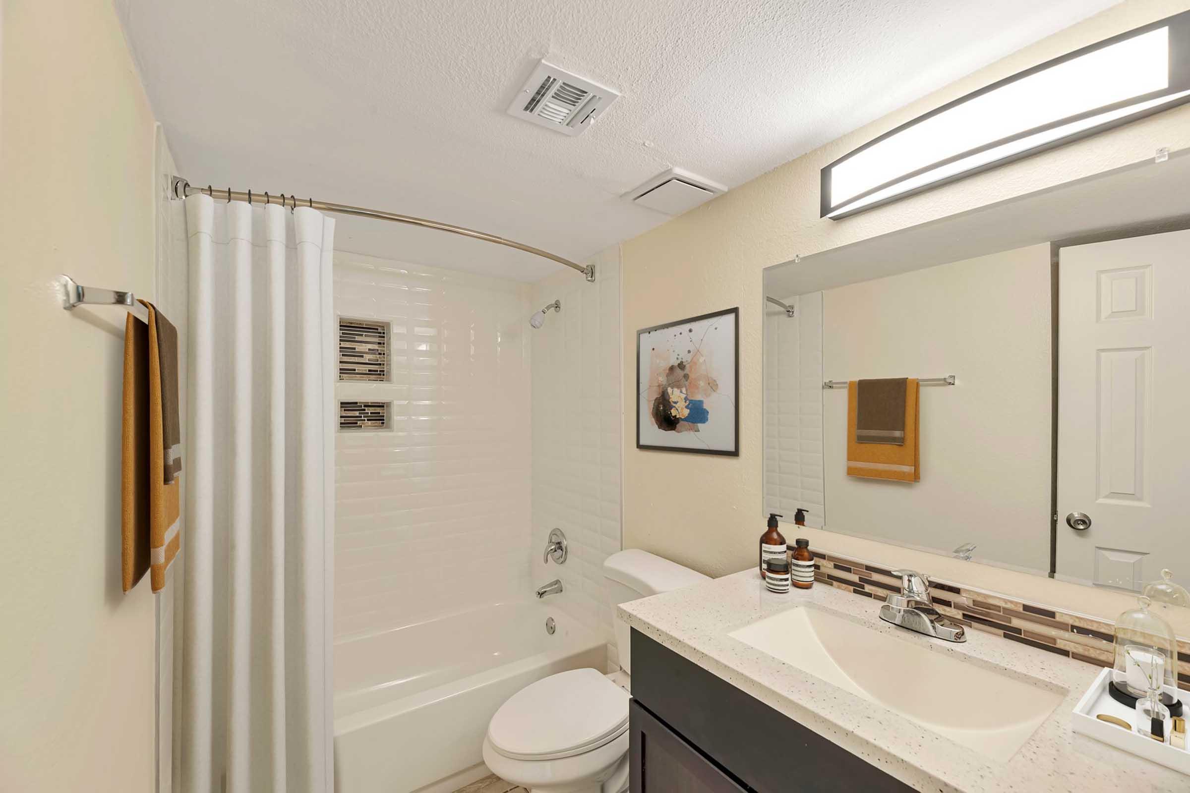 A modern bathroom featuring a white bathtub with a curved shower rod and a curtain. The walls are light-colored, and there’s a large mirror above a stylish sink with a dark cabinet. Decorative items and toiletries are neatly arranged on the countertop. A piece of artwork is displayed on the wall.