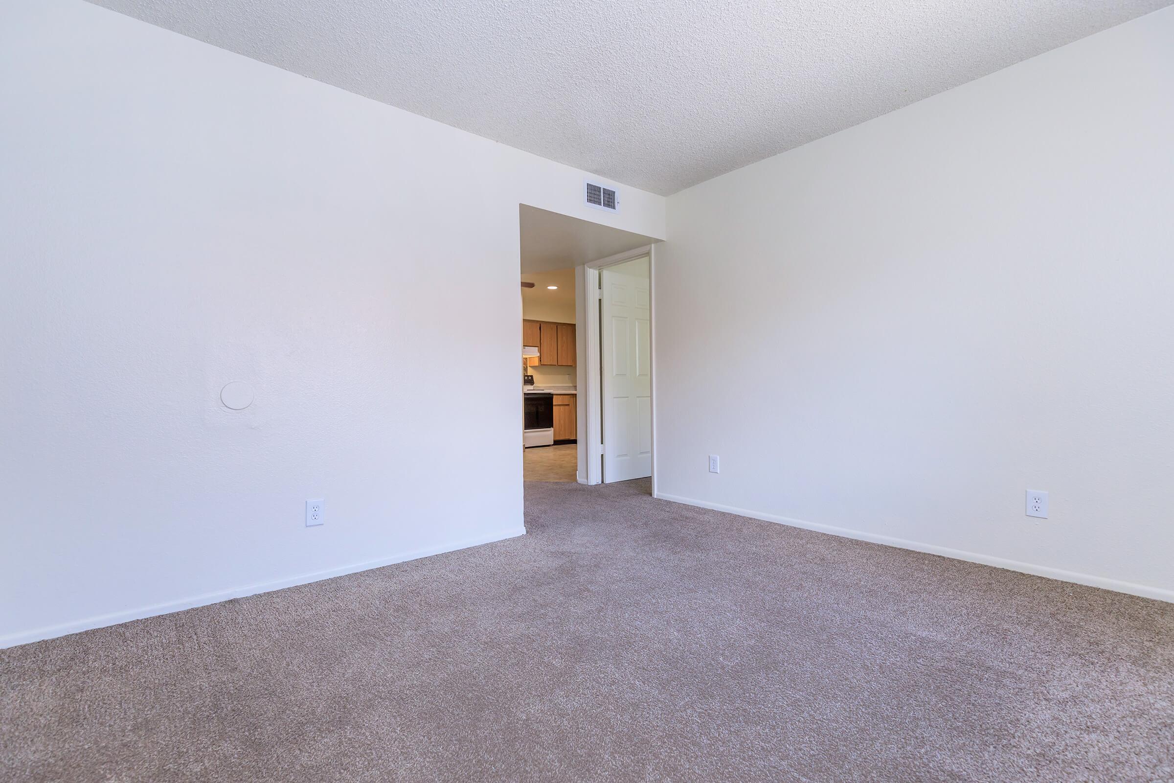 A spacious, empty room with light-colored walls and beige carpet. An open doorway leads to a kitchen area in the background, creating a sense of openness and light.