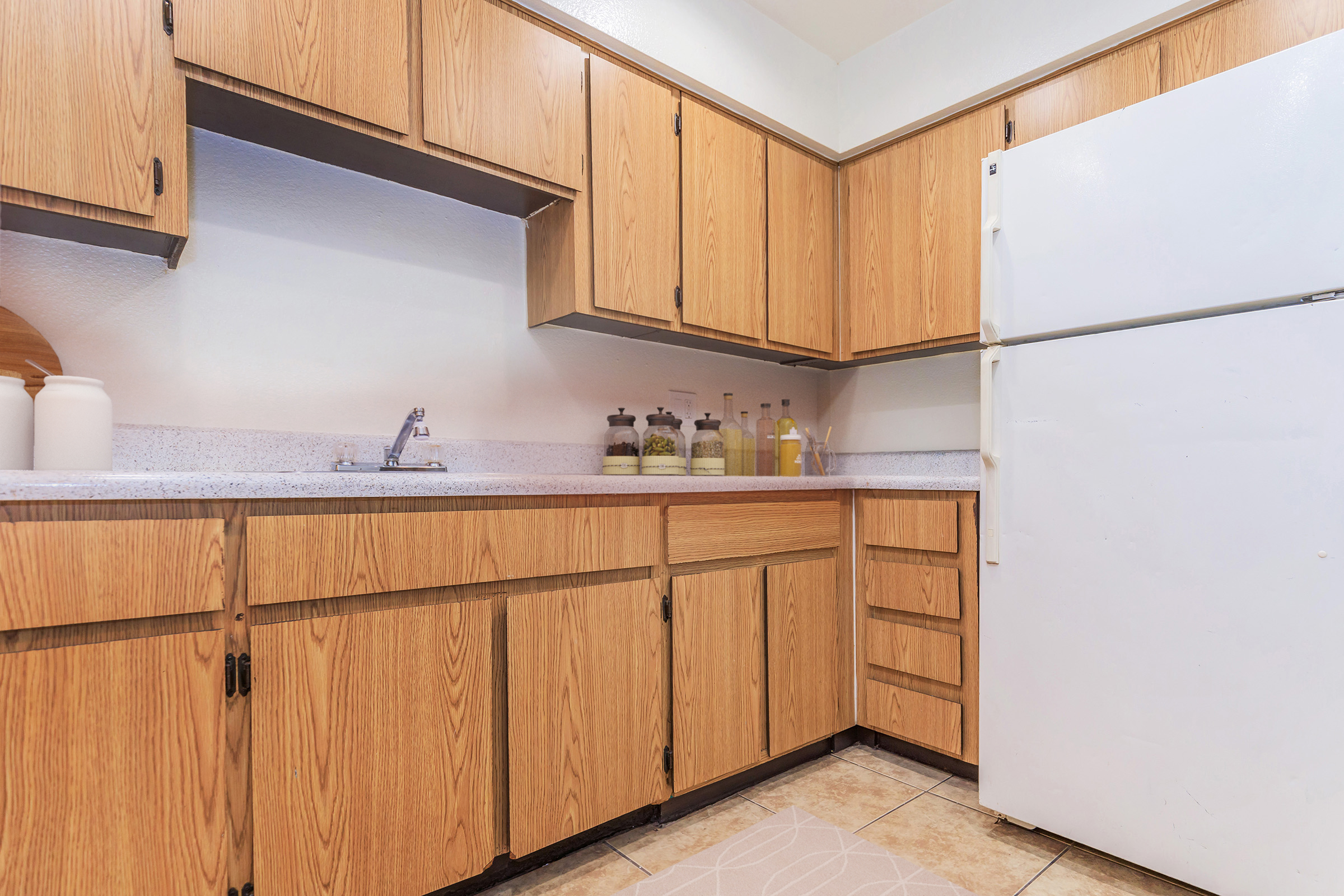 Modern kitchen with wooden cabinetry, a white countertop, and a stainless-steel sink. The cabinets are a light oak color, and there are several glass jars on the countertop. A refrigerator is visible on the right side. The floor features square tiles, and natural light illuminates the space.