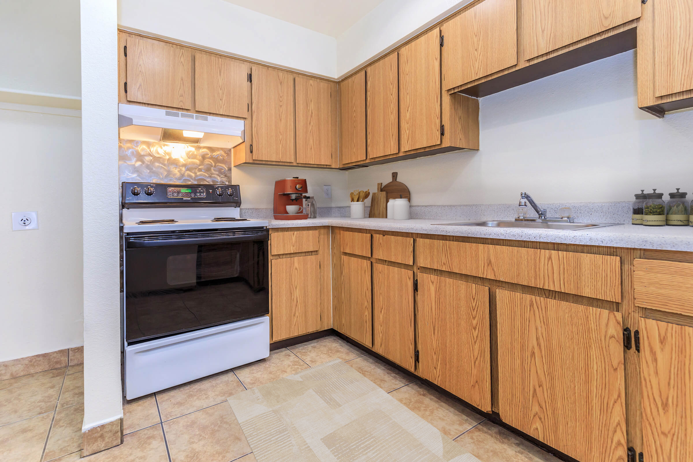 A kitchen with wooden cabinets, a white stove, and a countertop with a sink. There are kitchen appliances and utensils on the counter, and the floor is tiled. The overall design is functional and inviting, showcasing a clean and organized cooking space.