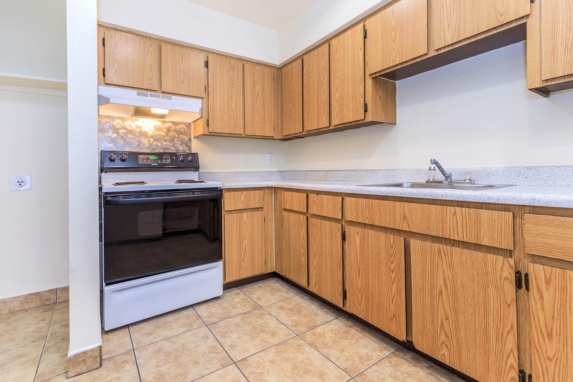A cozy kitchen featuring wooden cabinets, a white stove with an oven, and a stainless steel sink. The countertops are light-colored, and the floor is tiled. The kitchen has a clean and inviting atmosphere.