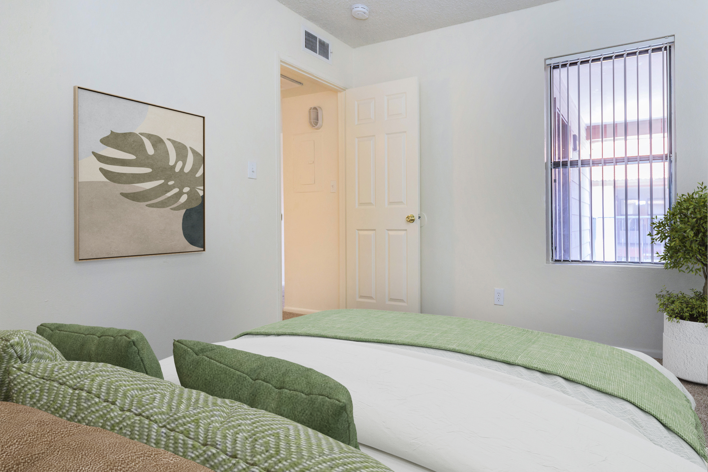 A cozy bedroom featuring a light green textured bedspread and several green accented pillows. A wall art piece with a leaf design is mounted above the bed. There is a closed door leading to another room and a window with vertical blinds allowing natural light into the space. A potted plant adds a touch of greenery.