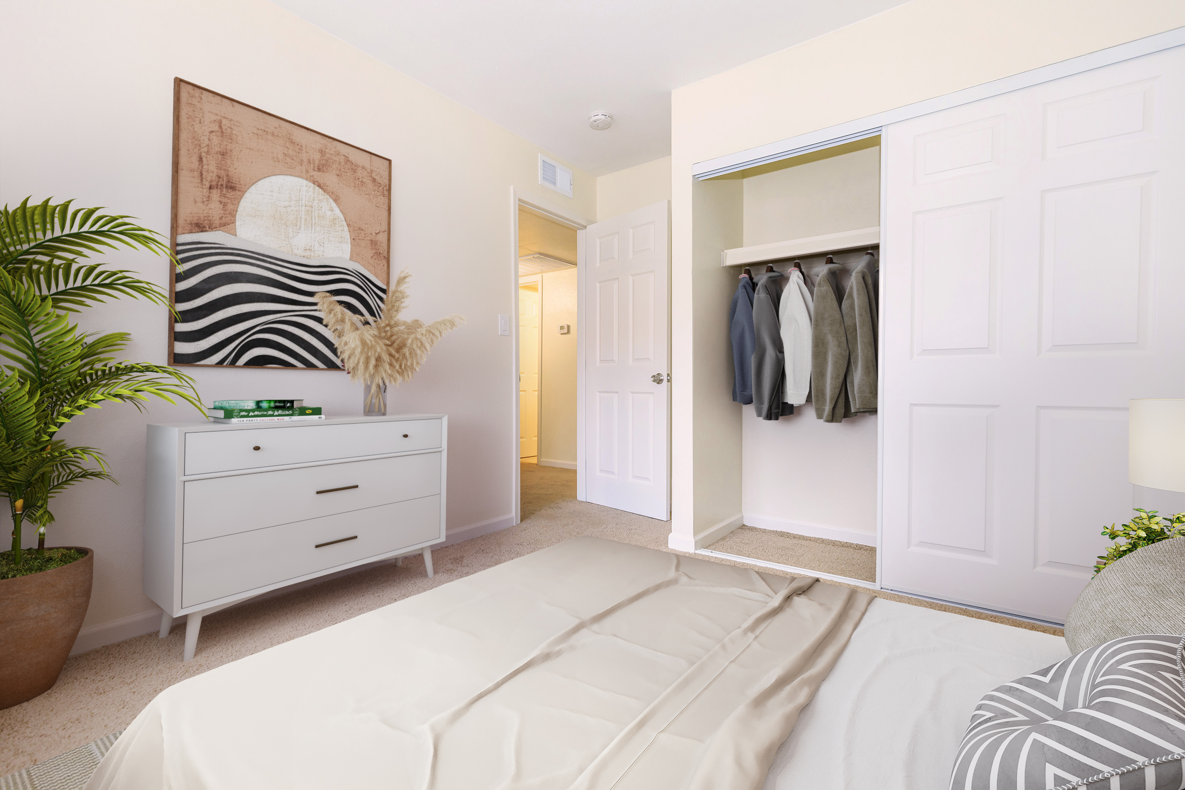 A cozy bedroom featuring a light-colored bedspread, a white dresser with plants and decor on top, and a closet with neatly hung suits. The room has subtle beige walls and a large piece of wall art depicting abstract waves. Natural light enters through a doorway, creating a warm atmosphere.