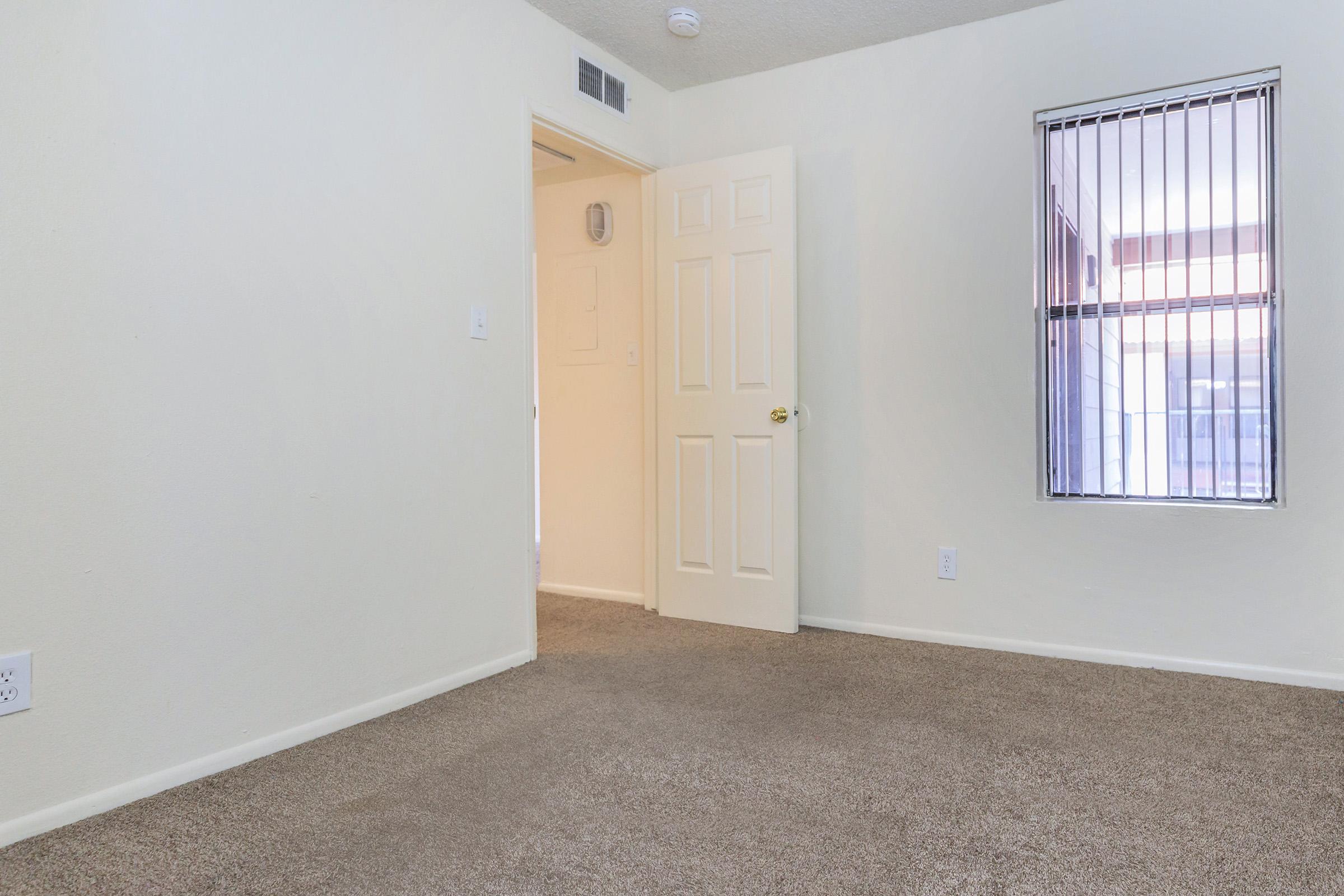 A small, empty room with light-colored walls and a beige carpet. A door is slightly open, leading to another space, and a window with vertical blinds lets in natural light. The room has a simple, clean look, suitable for various uses.