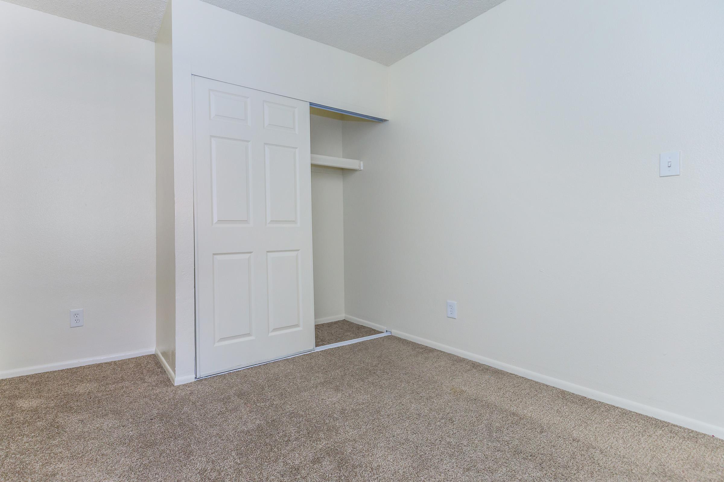 A minimal empty room featuring light-colored walls, a carpeted floor, and a closet with double doors. The closet has a shelf above, and there are two light switches on the wall. The overall atmosphere is neutral and bright, suitable for various uses or personal decoration.