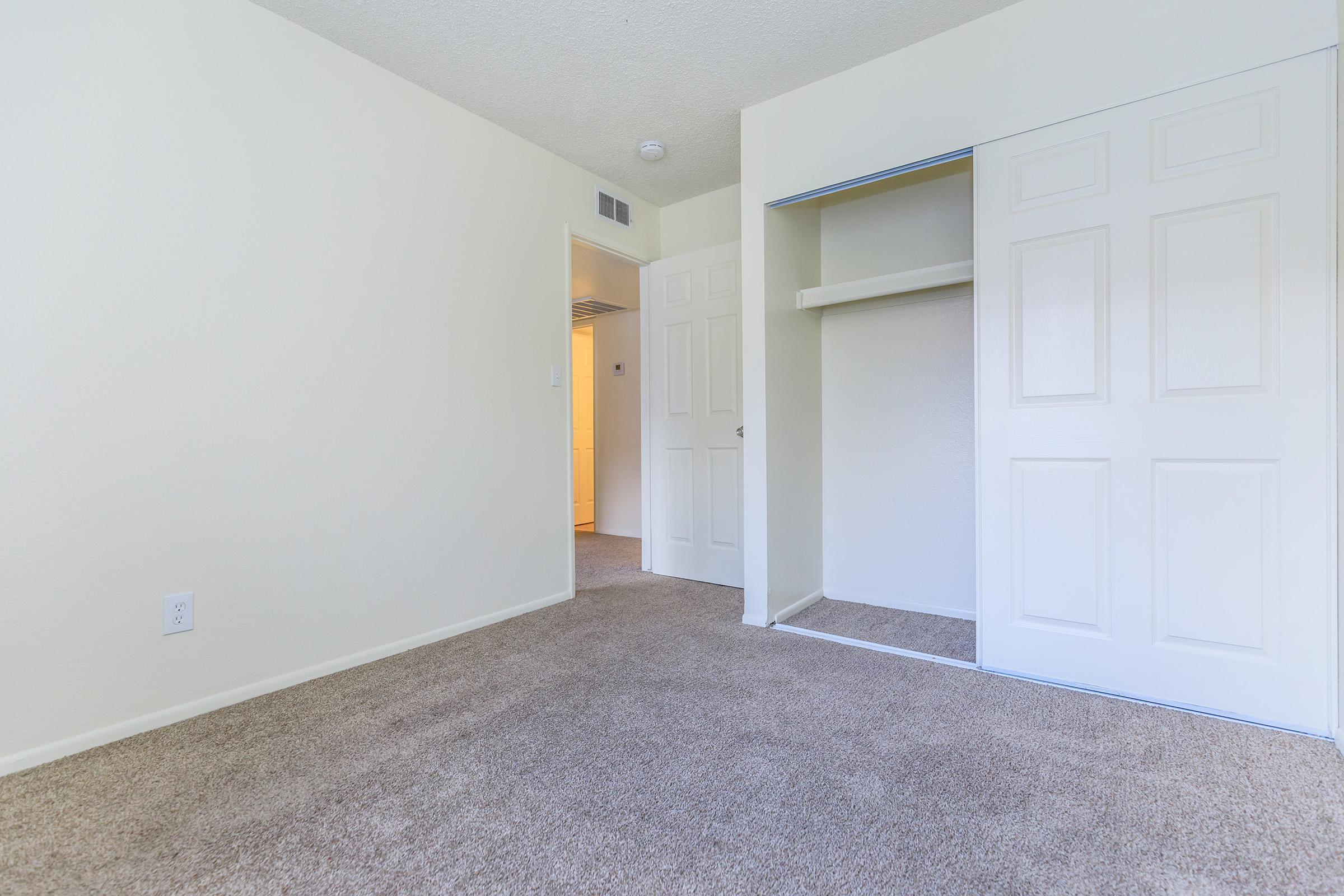 A spacious, empty room with light-colored walls and carpet flooring. There is a closed closet with double doors on one side, and a doorway leading to another room is visible in the background. The overall ambiance is bright and clean, ideal for personal decoration.