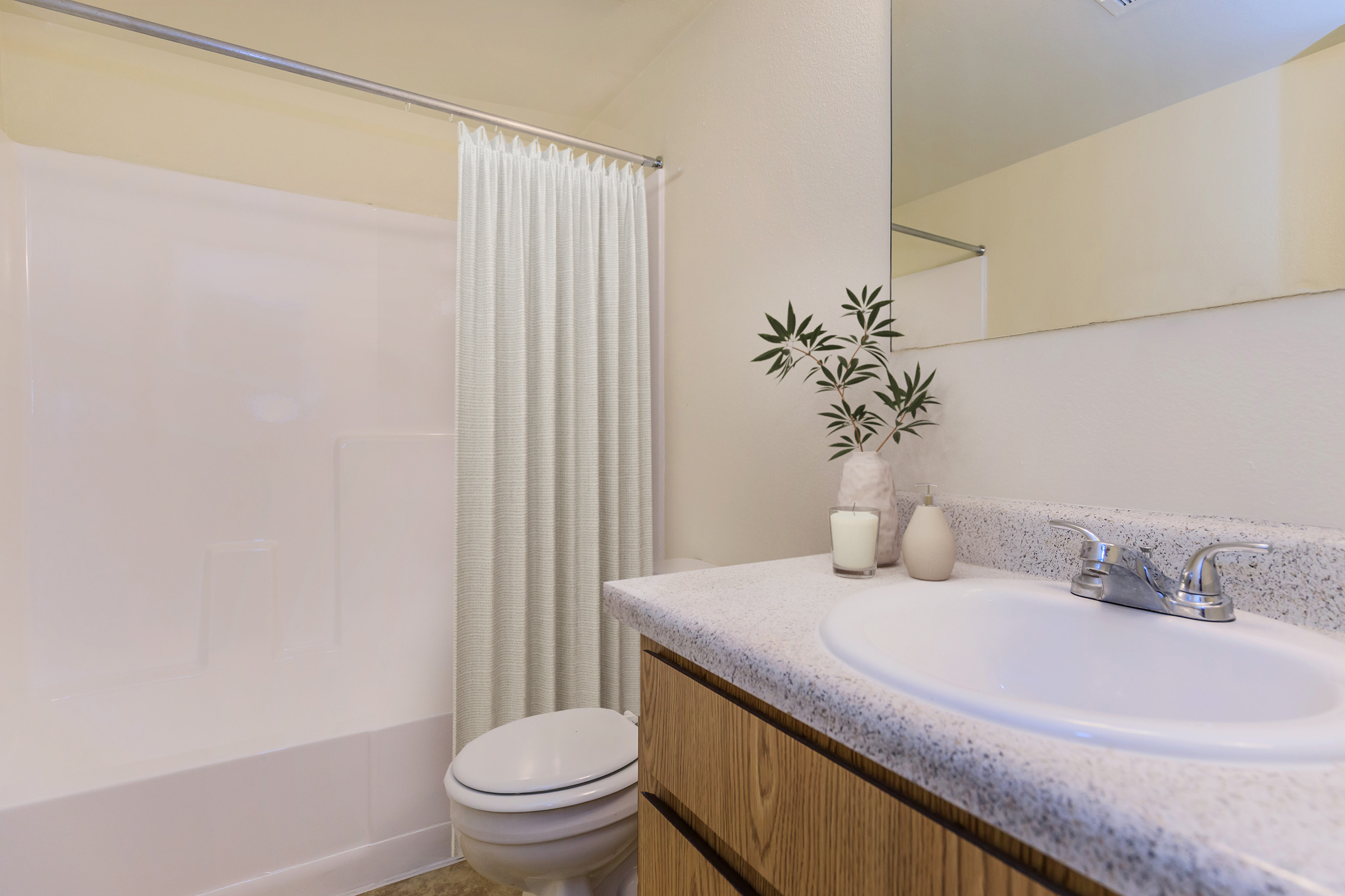 A clean bathroom featuring a white shower curtain, a bathtub with shower, a sink with a countertop, and a mirror. A small plant and candles are placed on the countertop, enhancing the warm and inviting ambiance of the space. Light-colored walls create a bright atmosphere.