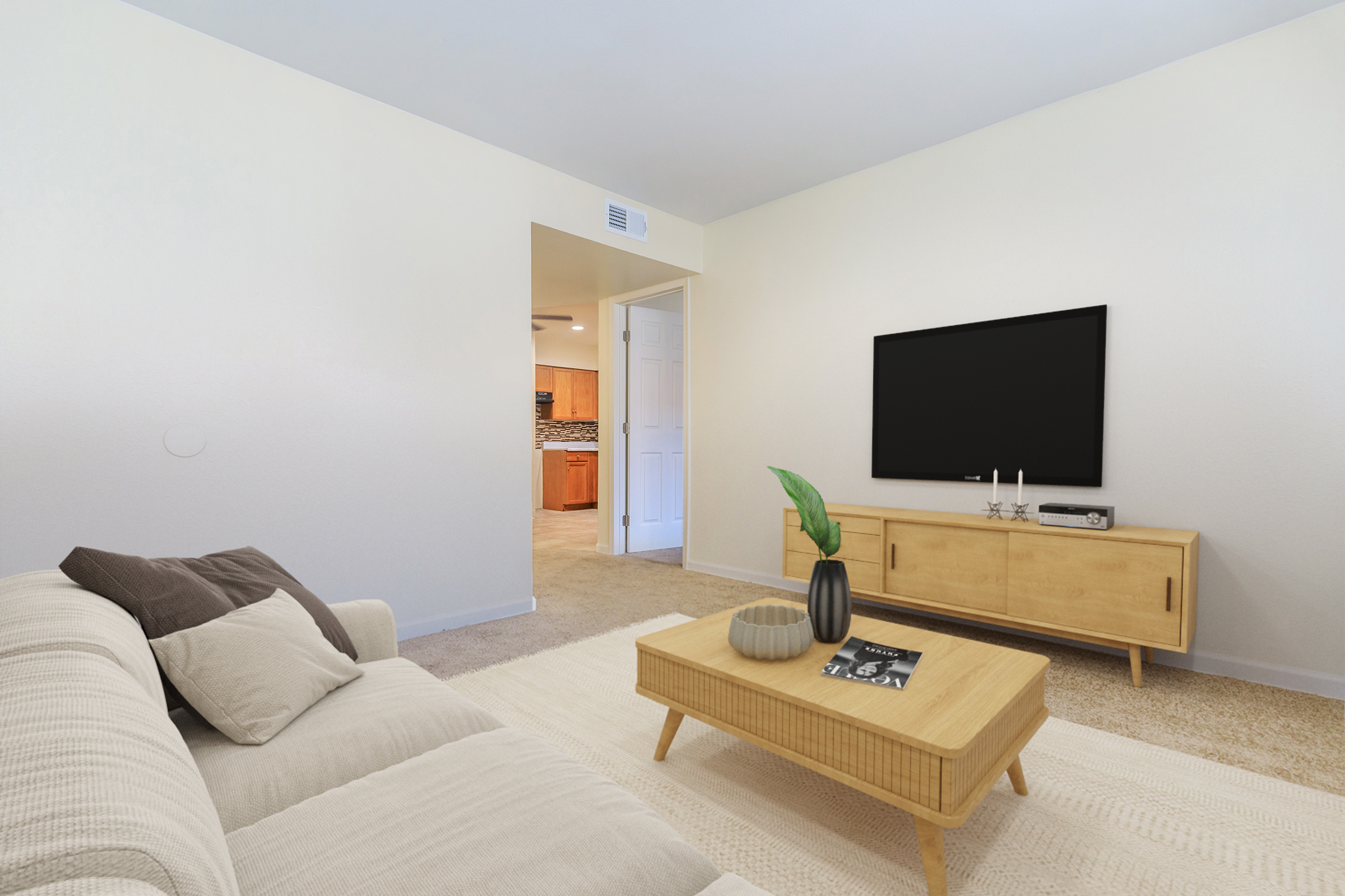 A cozy living room featuring a light-colored couch with throw pillows, a wooden coffee table with a decorative plant, and a modern media console with a large flat-screen TV. The space has a neutral color palette and connects to a kitchen visible in the background.