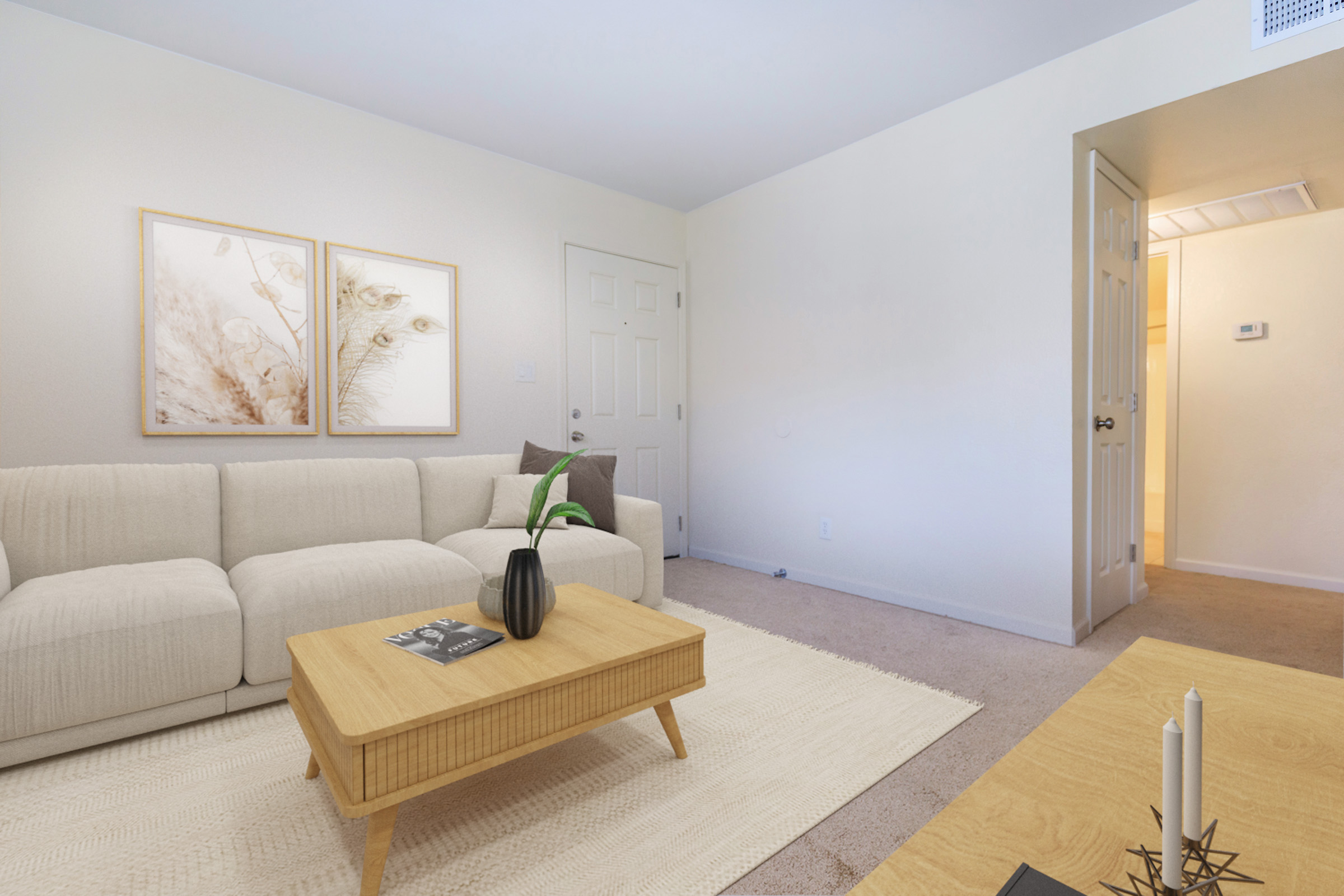 A cozy living room with a light-colored sofa, wooden coffee table, and decorative plant. Two framed artworks are hanging on the wall. The space is bright and inviting, with neutral tones and simple decor, leading to an open doorway seen in the background.