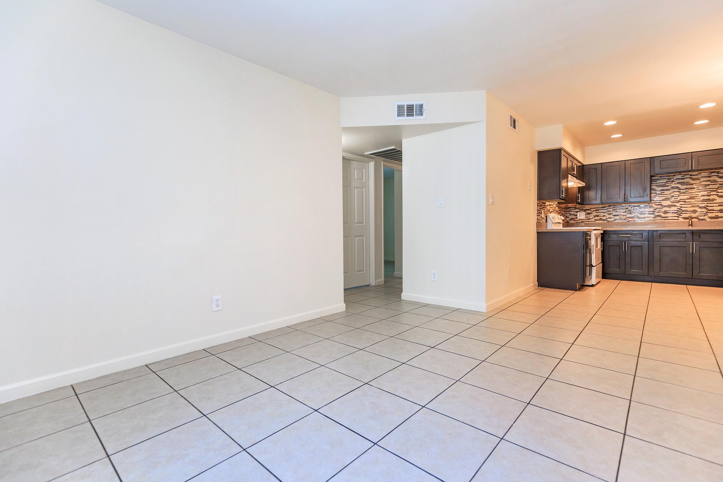Interior of a modern living space featuring a light-colored wall and tiled flooring. To the right, there's a kitchen area with dark cabinetry and a decorative backsplash. The room is well-lit, showcasing an open floor plan, with a doorway leading to another room visible in the background.