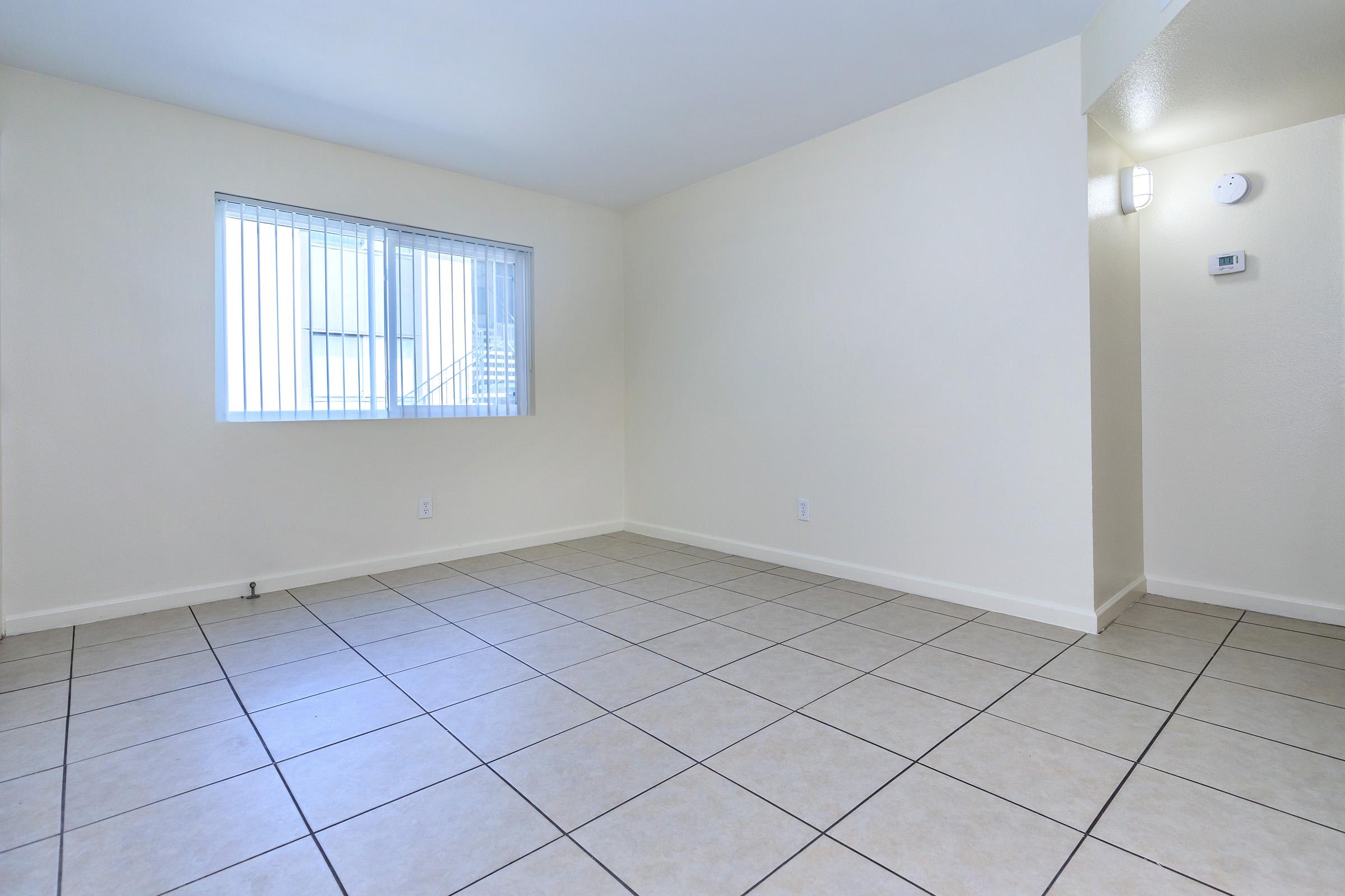 Empty room with beige walls and tiled flooring. There is a window with vertical blinds on one wall, allowing natural light to enter. The opposite wall features a light fixture and a thermostat. The space is clean and spacious, suitable for various purposes.