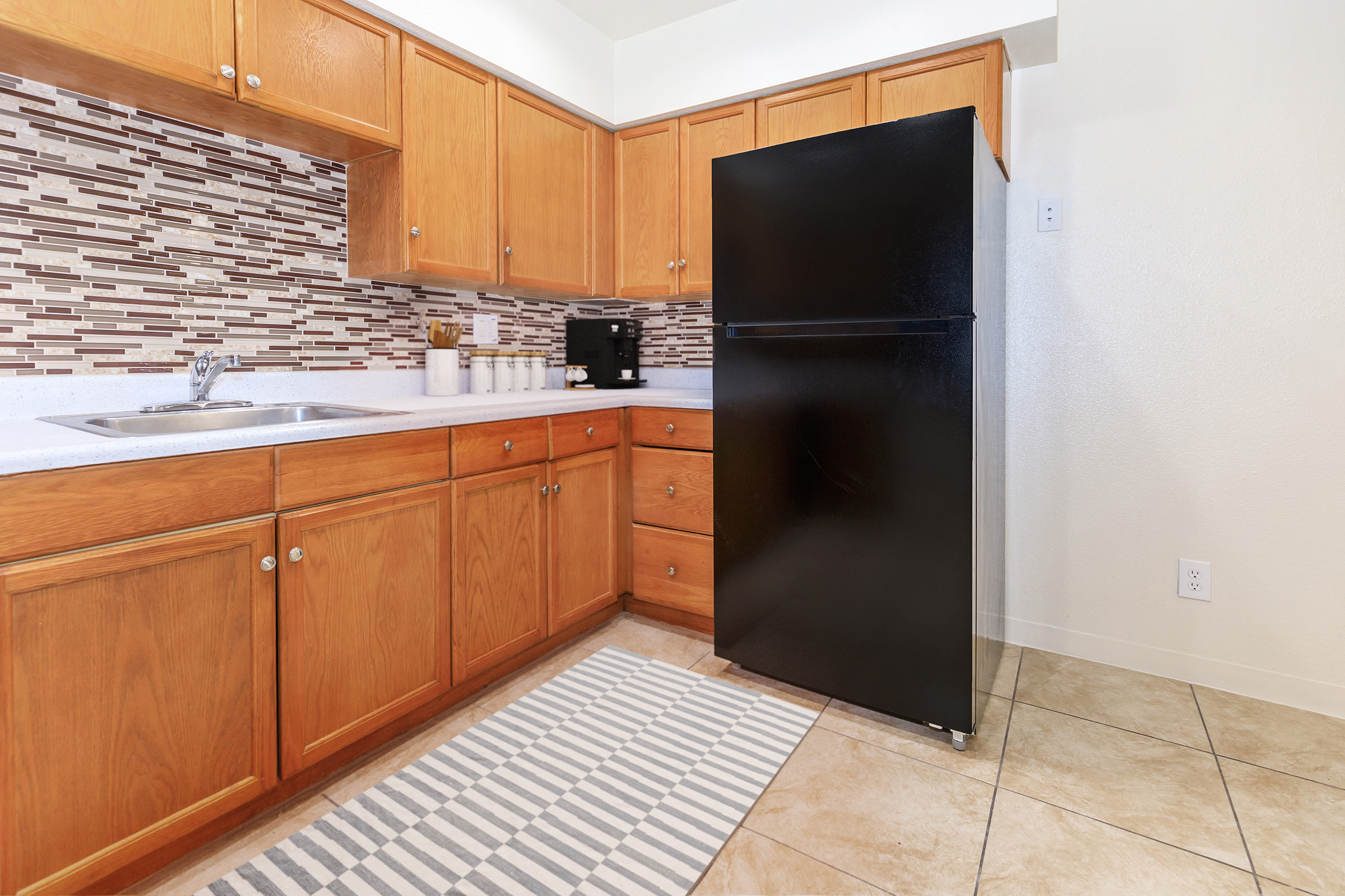 A modern kitchen with wooden cabinets, a black refrigerator, and a stainless steel sink. There is a decorative backsplash made of small tiles. A striped rug is positioned on the tiled floor. The space is well-lit and organized, featuring a coffee maker on the countertop.