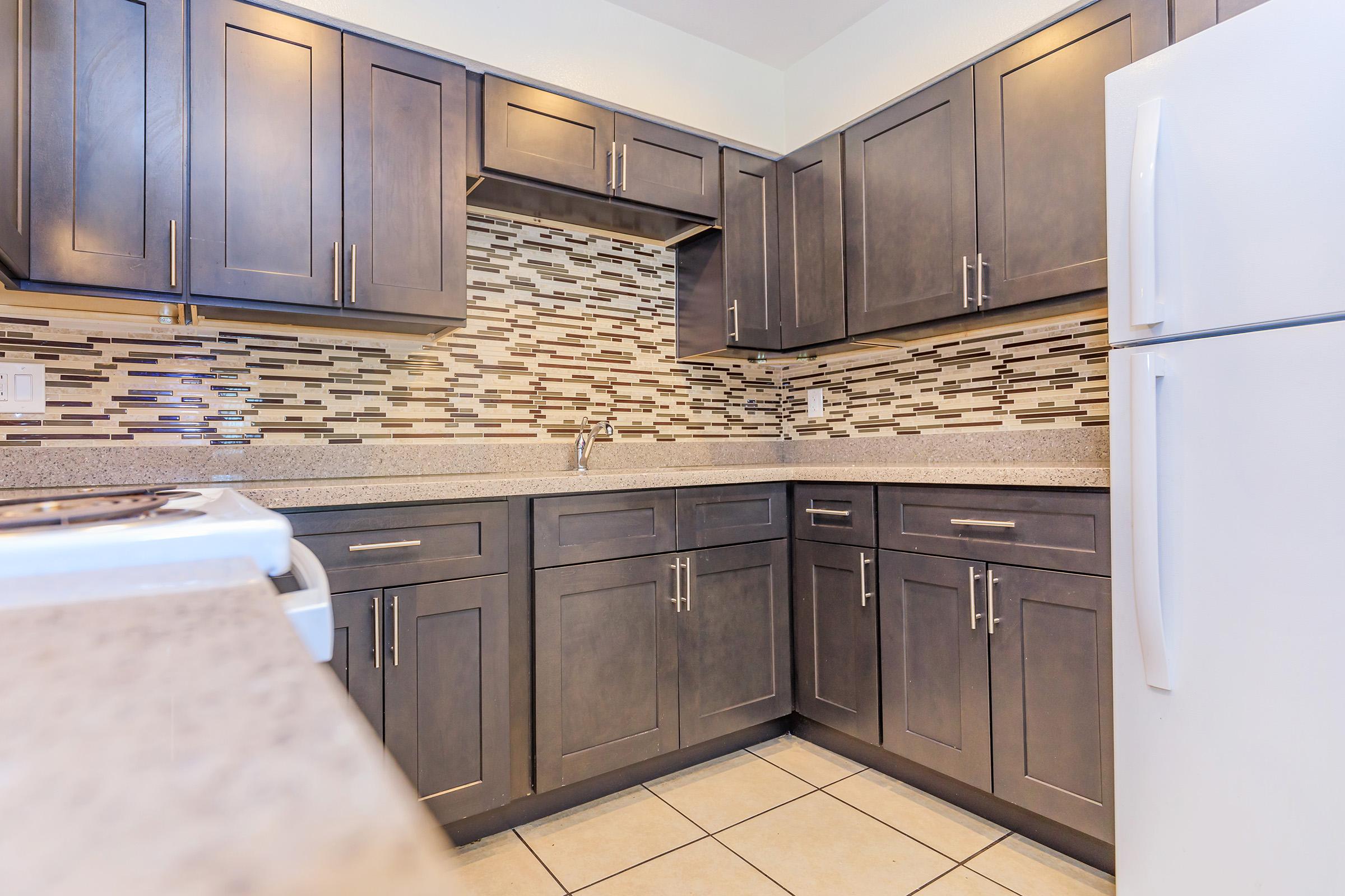 A modern kitchen featuring dark wooden cabinets, a decorative tiled backsplash, and a light-colored countertop. The space includes a stainless steel sink, and a white refrigerator is visible in the corner. The floor consists of beige tiles, creating a sleek and contemporary appearance.