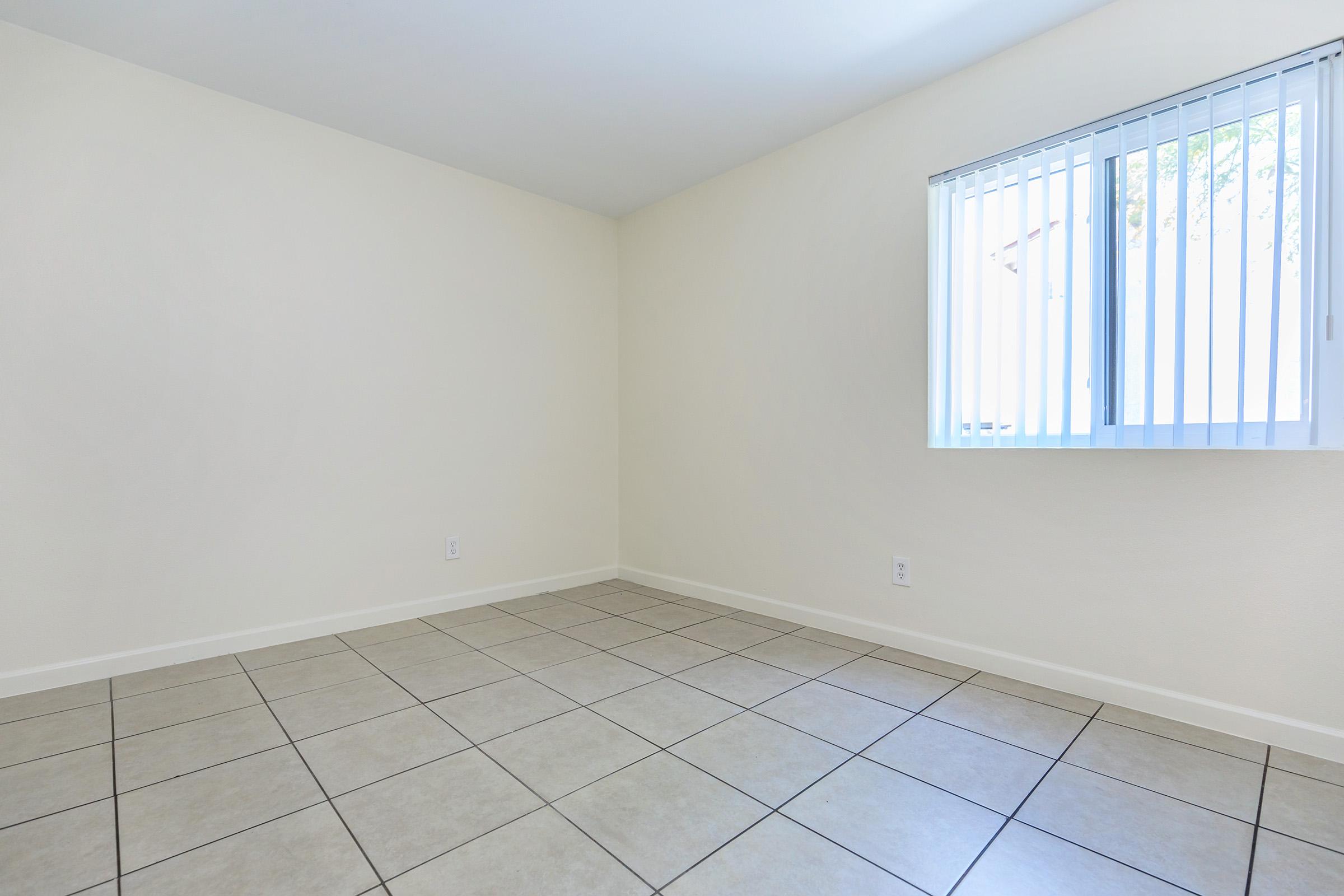 A vacant room with light-colored walls and tiled flooring. There is a window with horizontal blinds allowing natural light to enter the space. The room is empty, creating a sense of openness and potential for various uses or furnishings.