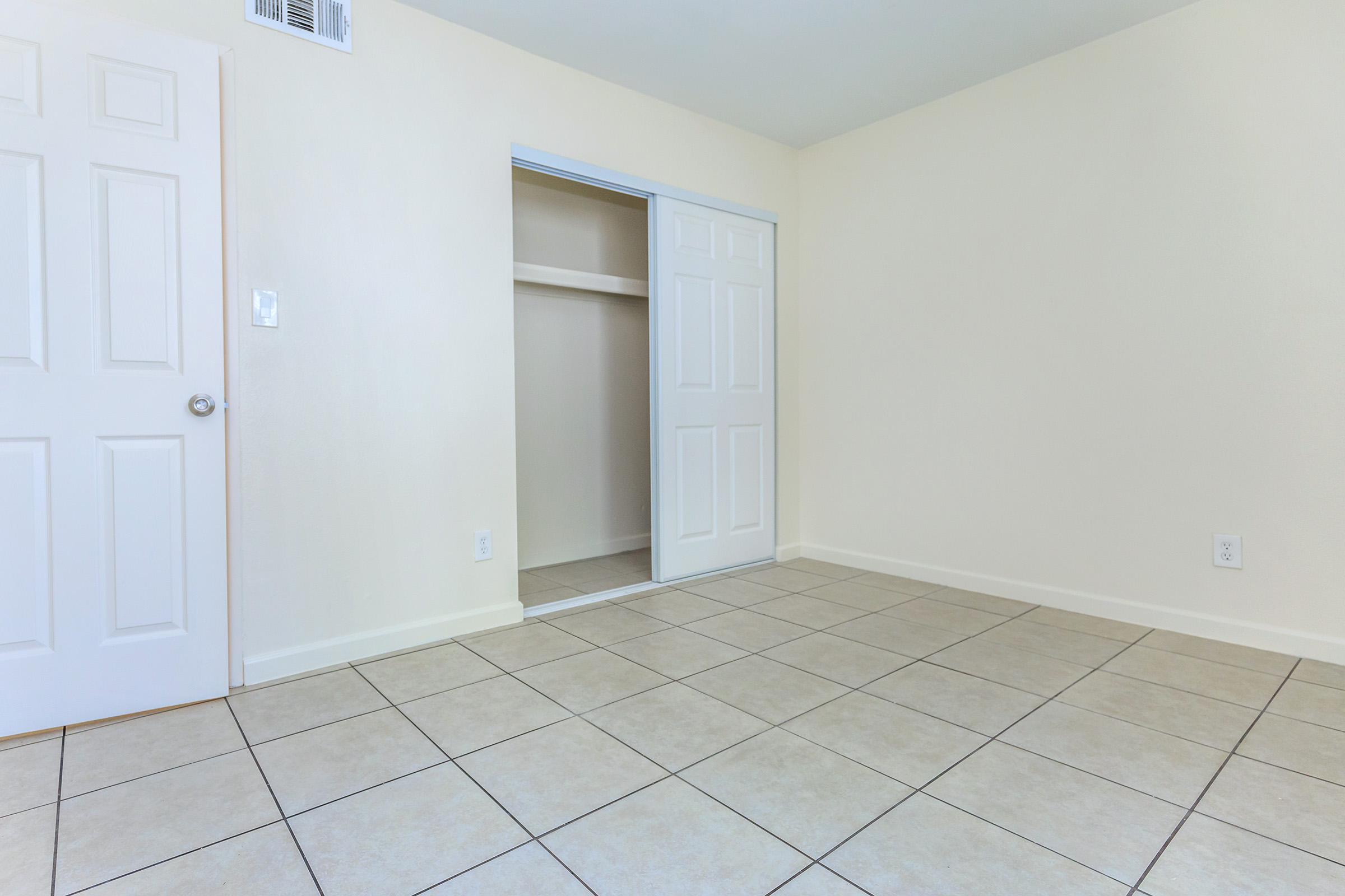 An empty room with light-colored walls and a tiled floor. The space features a closet with double doors open to reveal the interior. There is a single door on the left side, leading to another area. Natural light illuminates the room, creating a bright and spacious feel.