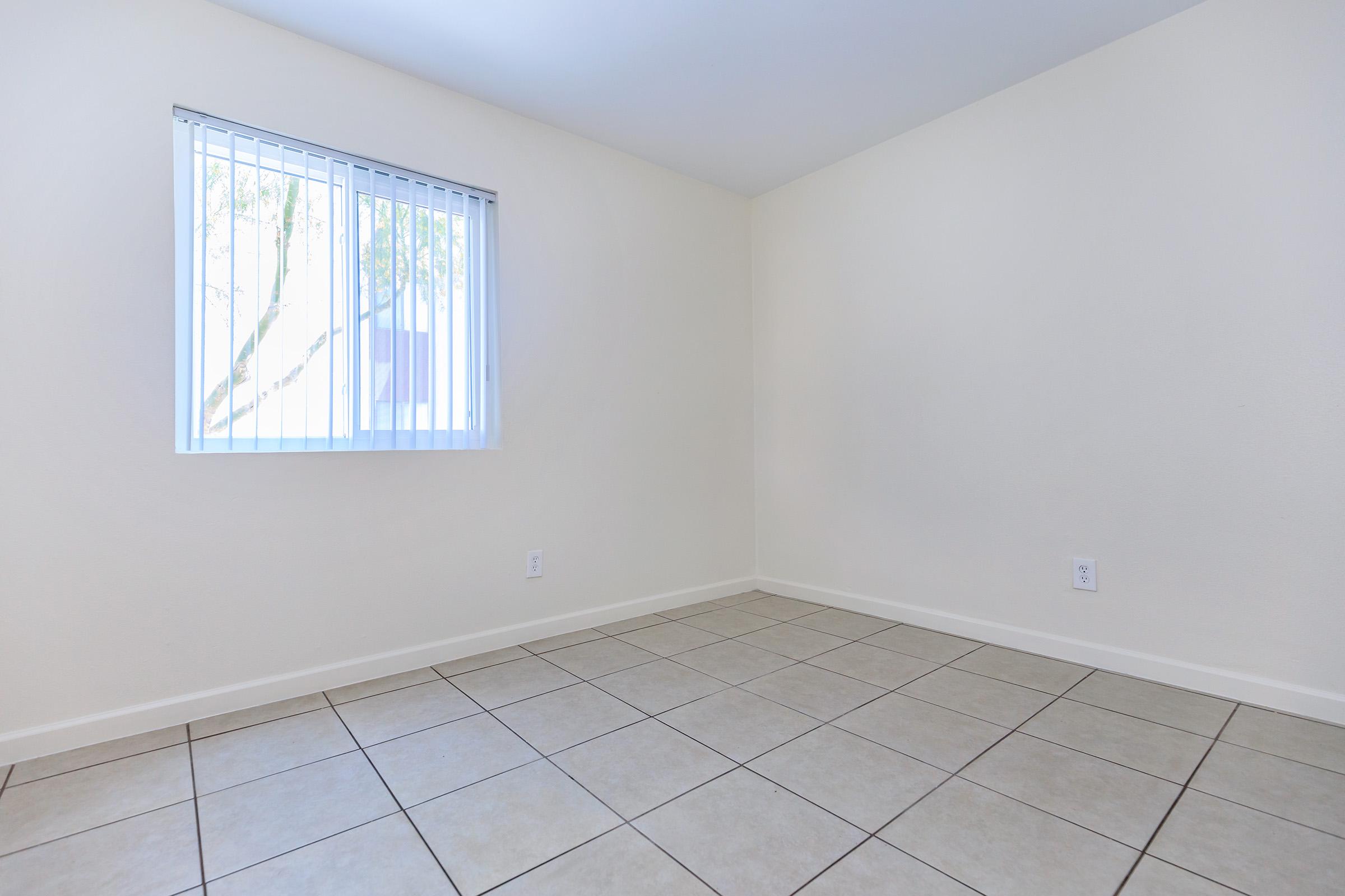 An empty room with light beige walls and large square tiles on the floor. There is a window with vertical blinds allowing natural light to enter, and a view of a tree outside. The space is uncluttered and minimal, ideal for various uses.