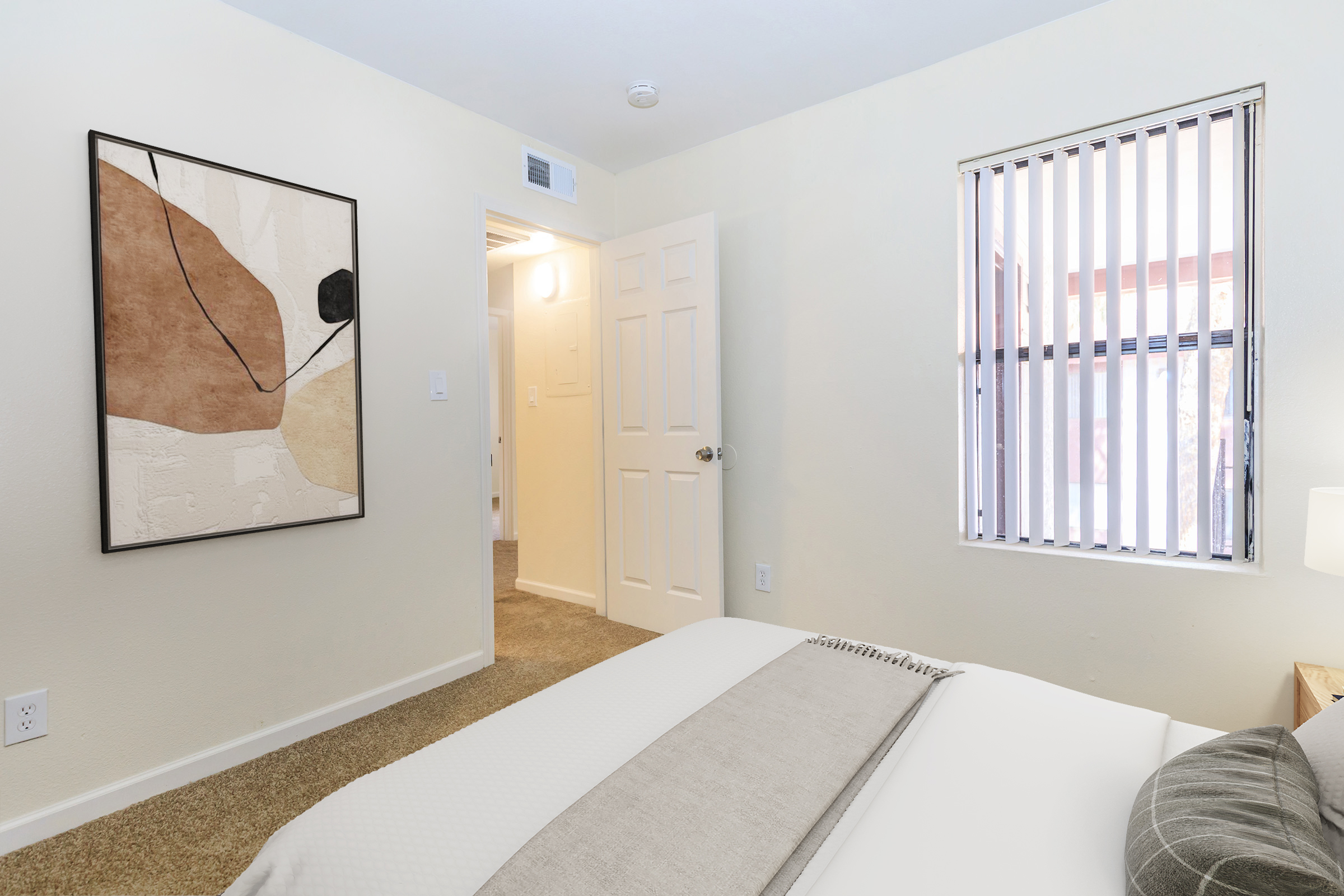 A cozy bedroom featuring a bed with a light gray blanket, minimalist artwork on the wall, and a window with vertical blinds. The room has warm beige walls and a plush carpet, creating a comfortable and inviting atmosphere. A door leads to another room, visible in the background.