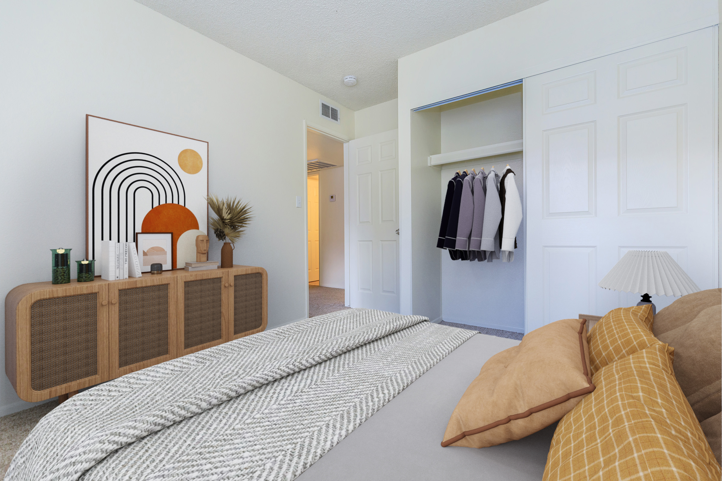 Interior of a modern, well-lit bedroom featuring a bed with decorative pillows, a stylish dresser with decor items, and a closet with hanging clothes. A large artwork with abstract shapes is on the wall, and a small lamp is on the nightstand. The room has a clean and minimalist aesthetic.