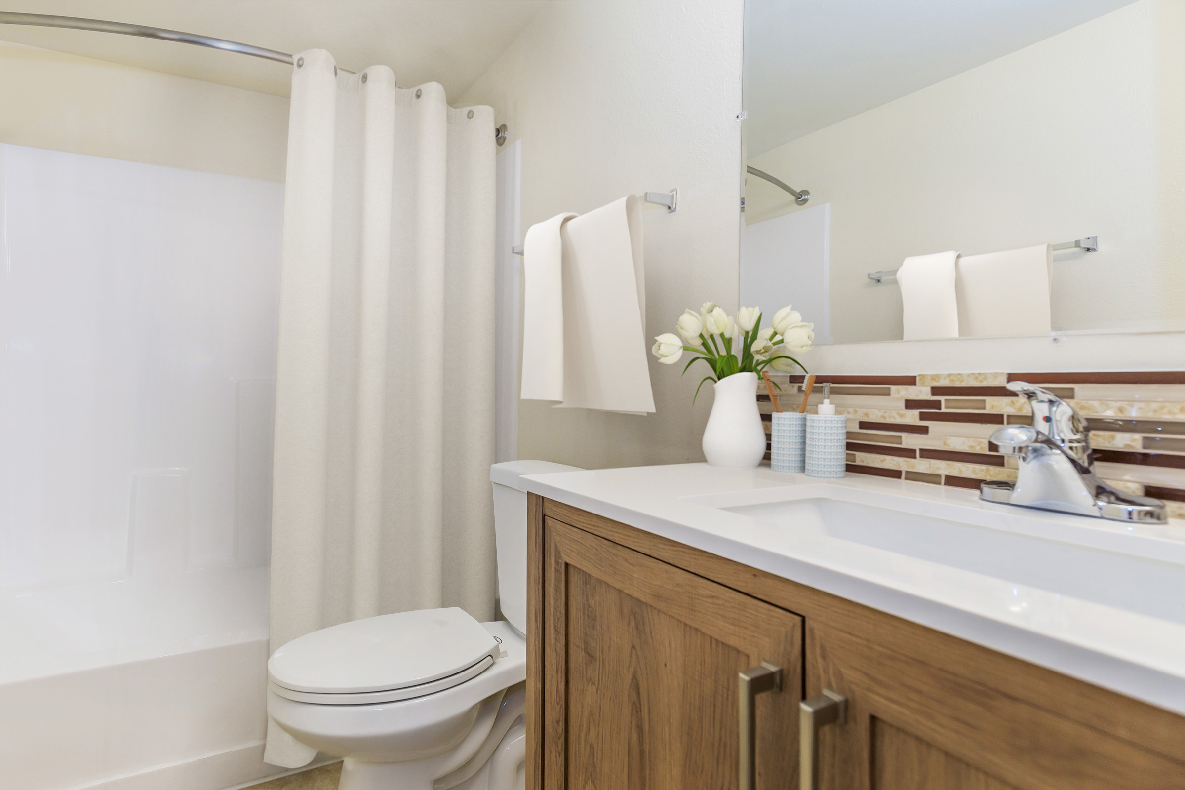 A modern bathroom featuring a shower with a white curtain, a vanity with wooden cabinetry, a sink with a silver faucet, and decorative elements like a white vase with tulips and a container on the counter. The walls are light-colored, creating a bright and airy ambiance.