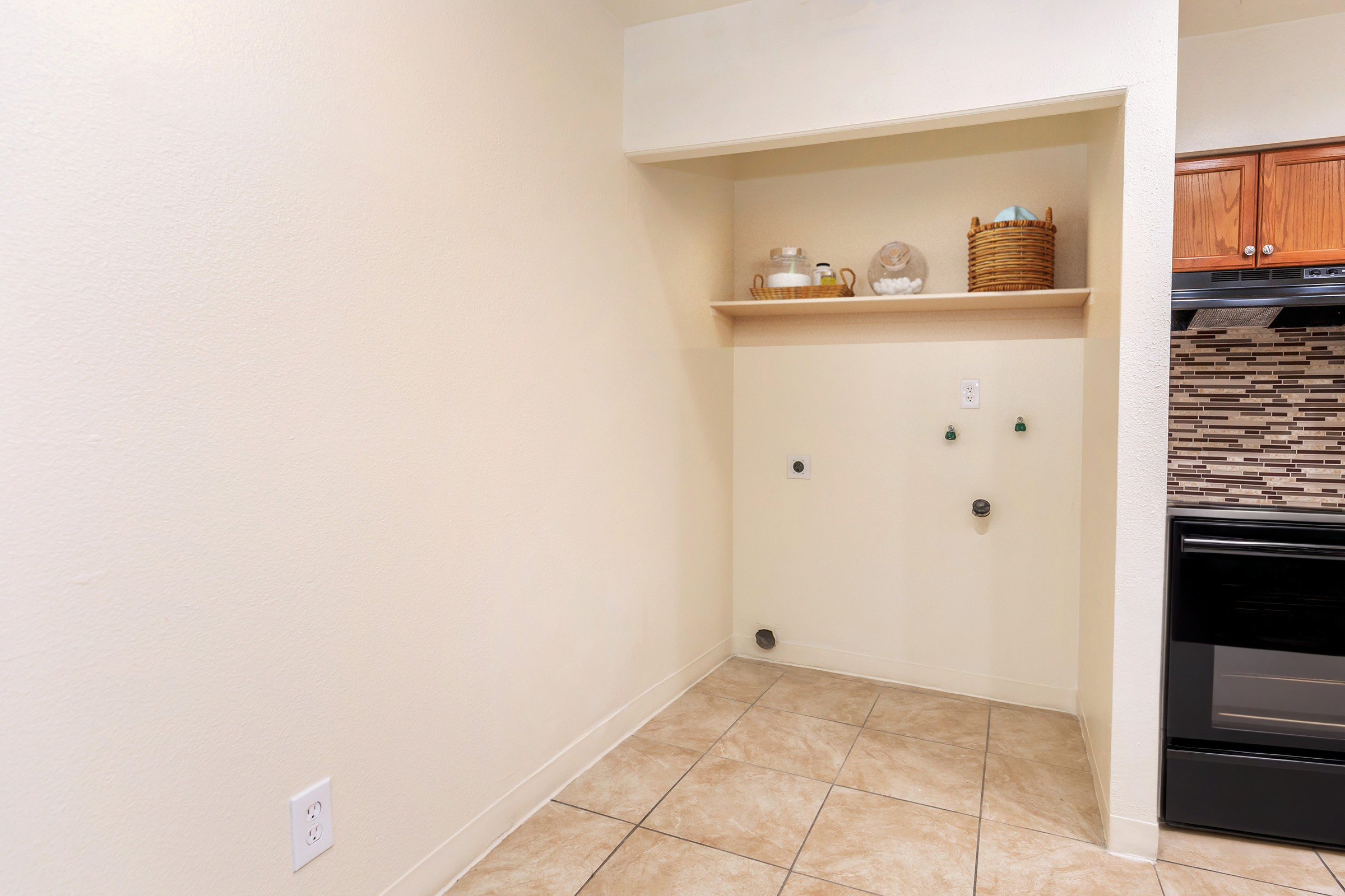Empty kitchen space with beige walls and tiled floor. A wooden shelf is mounted on the wall, holding decorative items. Below the shelf, there are two water connections and a wall outlet. A black stove is visible on the right, contrasting with the light-colored walls and floor tiles.