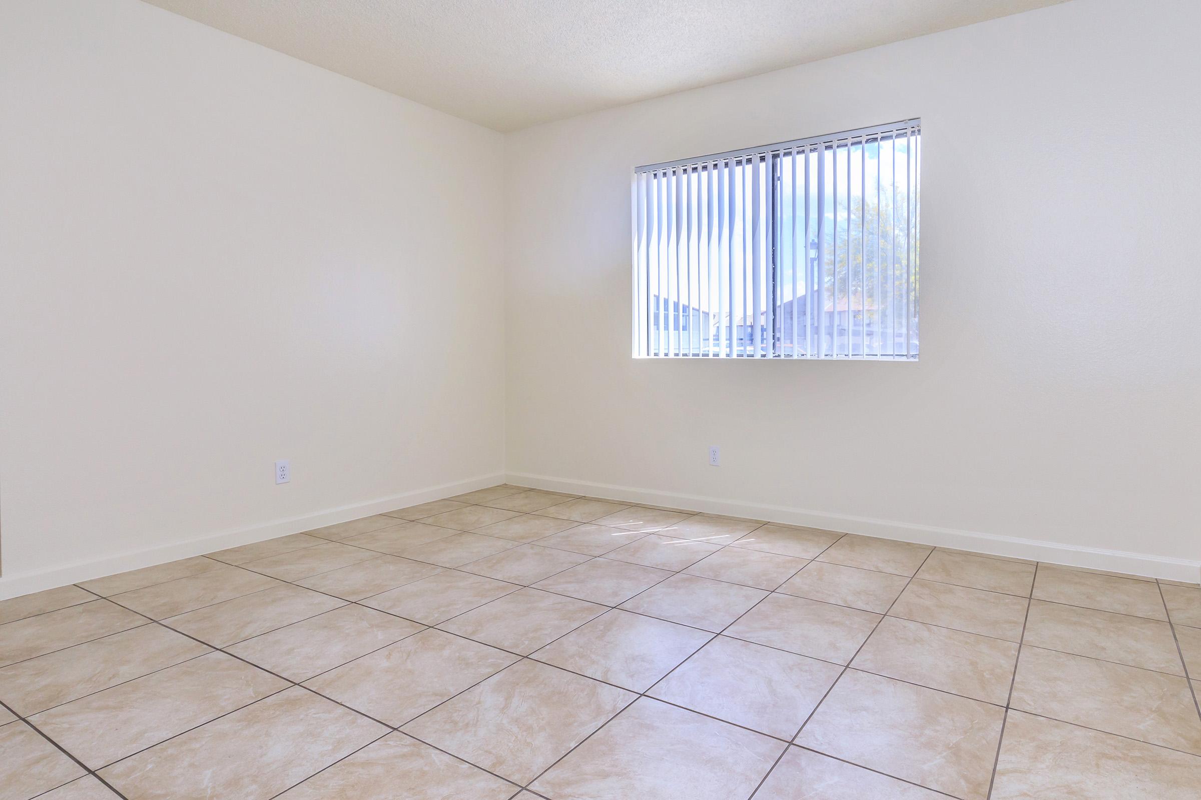 A spacious, empty room with light-colored walls and a tiled floor. A window with vertical blinds allows natural light to enter, illuminating the clean and minimalist space. The room has no furniture, creating an open and airy atmosphere.