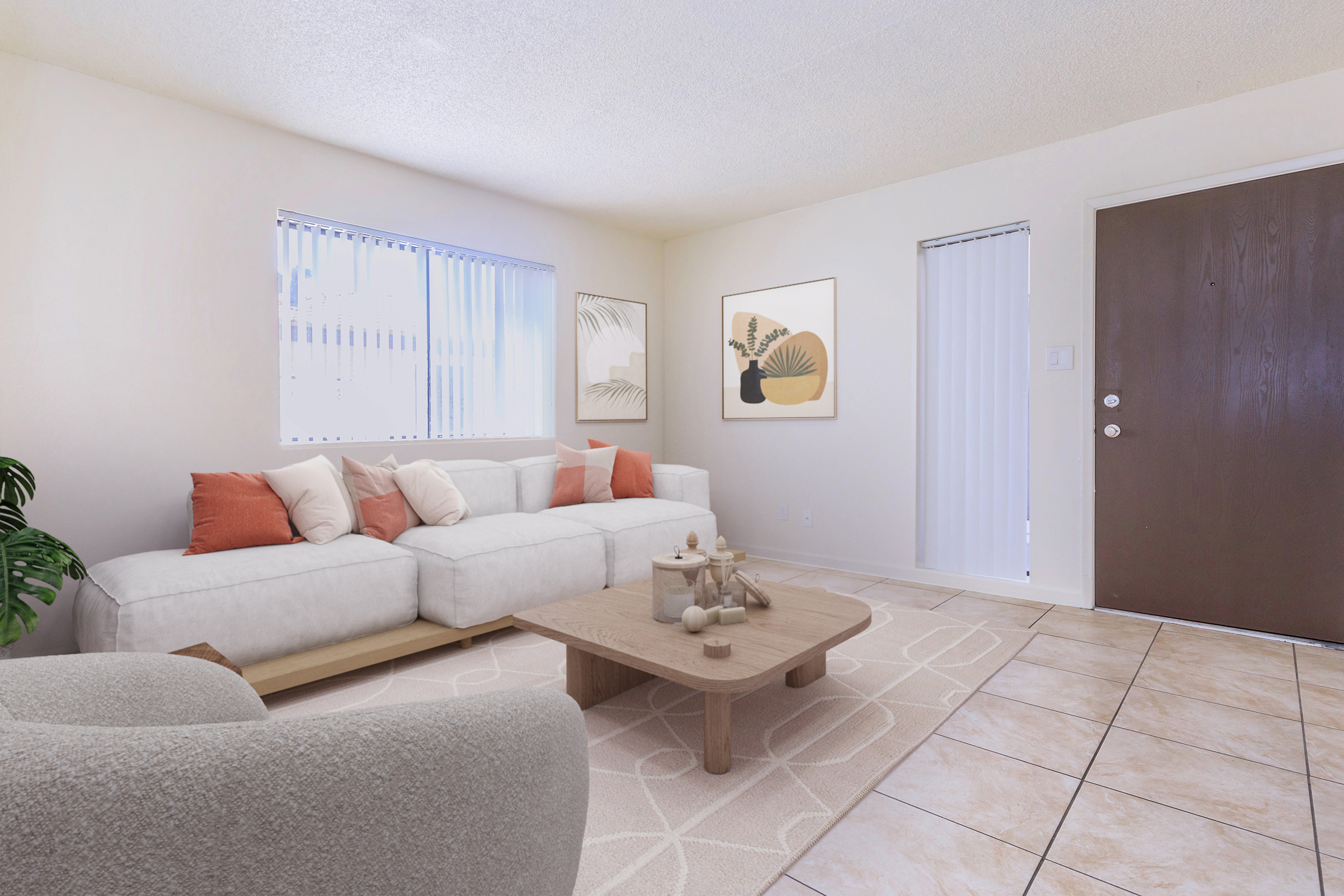 A bright and airy living room featuring a white sectional sofa with orange and cream pillows, a wooden coffee table with decorative items, and light-colored tile flooring. The walls are adorned with minimalist artwork, and large windows allow natural light to fill the space. A front door is visible in the background.