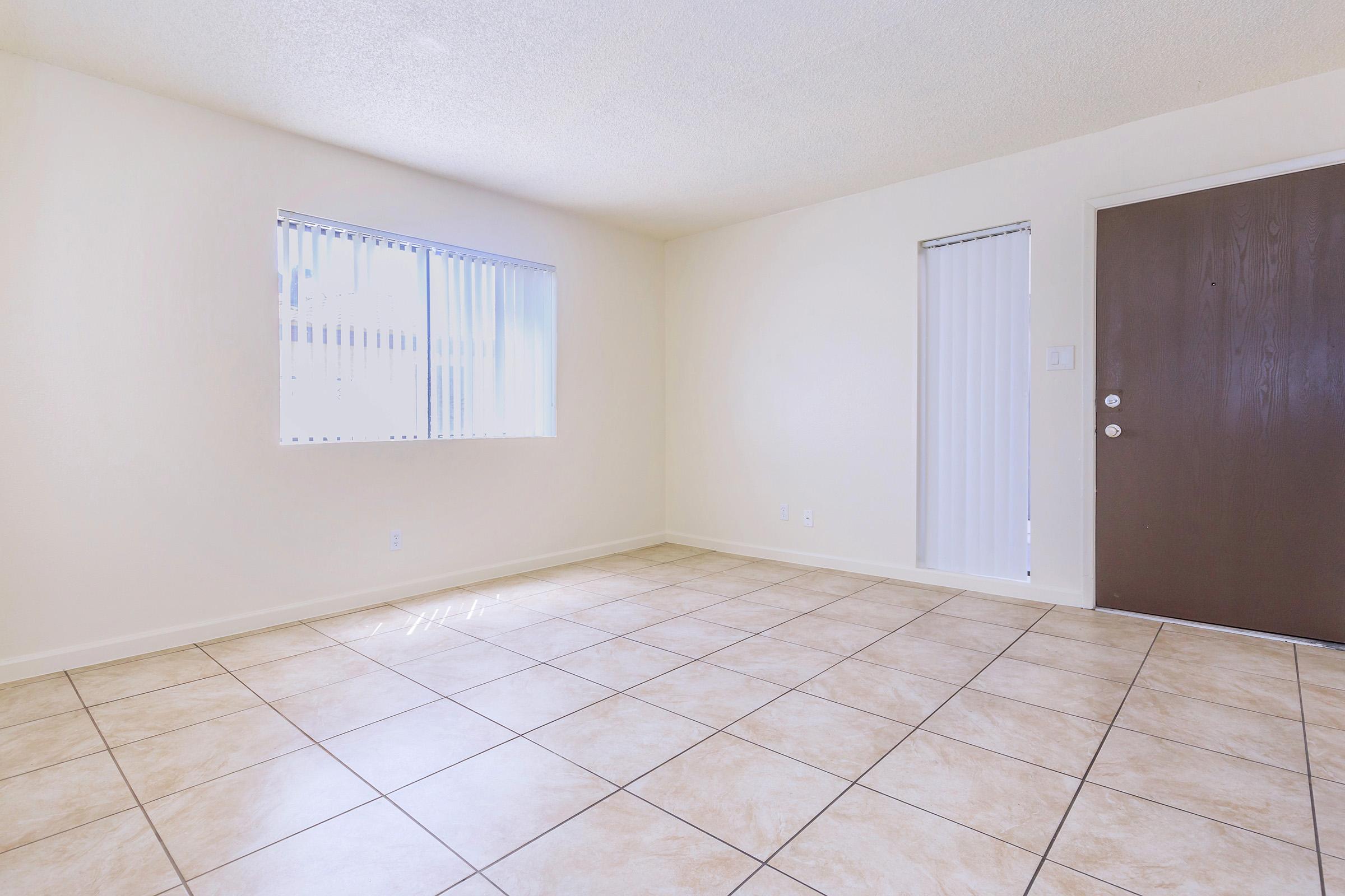 A bright and empty room with light-colored walls and tiled flooring. There are two windows allowing natural light, and a brown door on the right side. The space is uncluttered and ready for furnishing.
