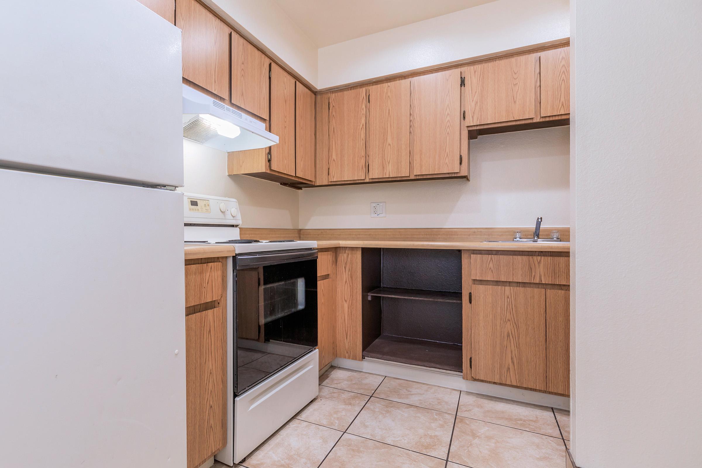 A compact kitchen featuring wooden cabinetry, a white stove and oven, a refrigerator, and a small sink area. The tiled floor complements the rustic style, and there is an open shelf space in one of the cabinets. The overall design is functional and simple, suitable for small living spaces.