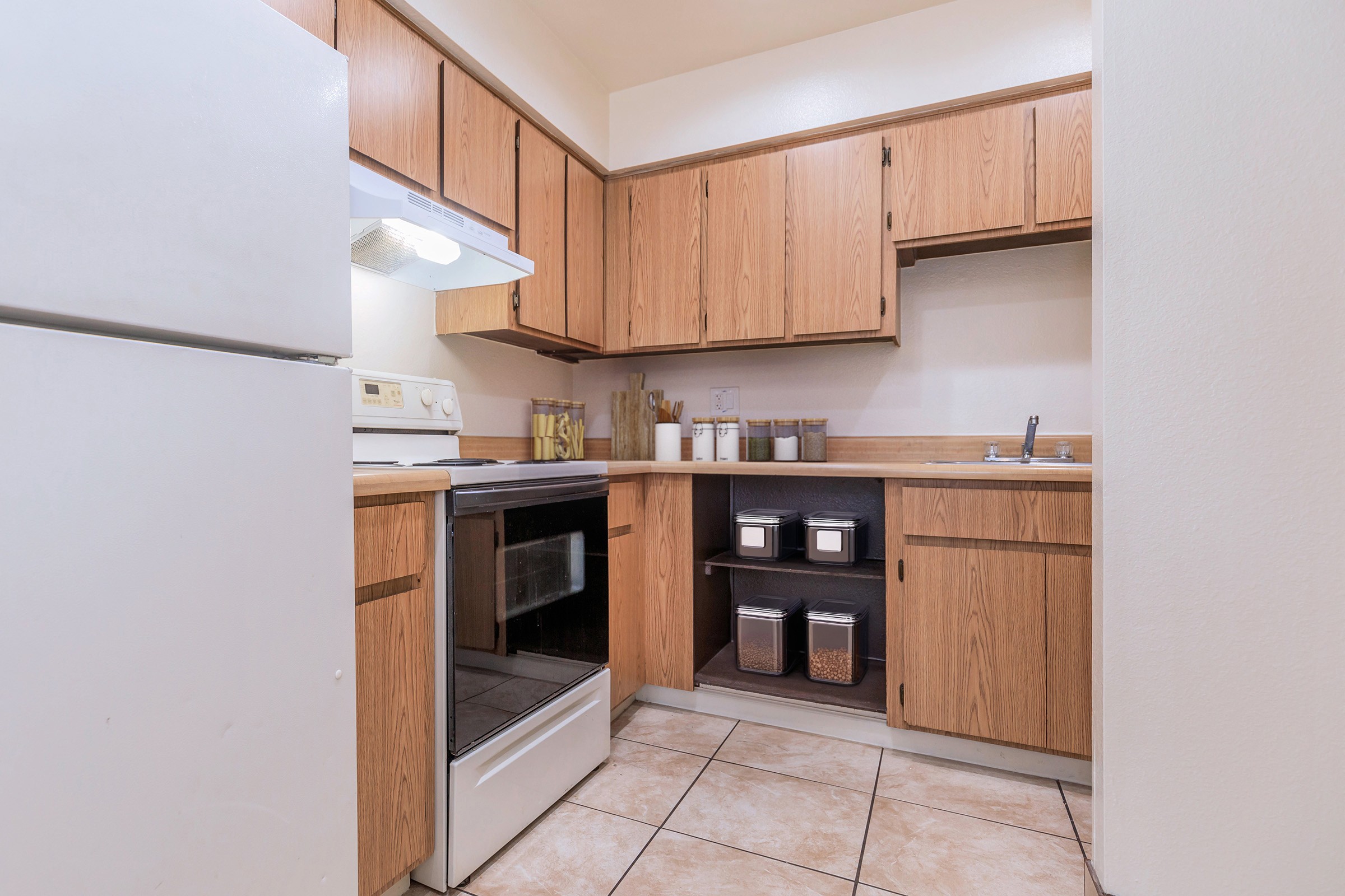 A cozy kitchen featuring wooden cabinets, a white stove, and an oven. There's a refrigerator on the left, and the countertop is organized with containers and kitchen utensils. The tiles on the floor are light-colored, creating a warm and inviting atmosphere.