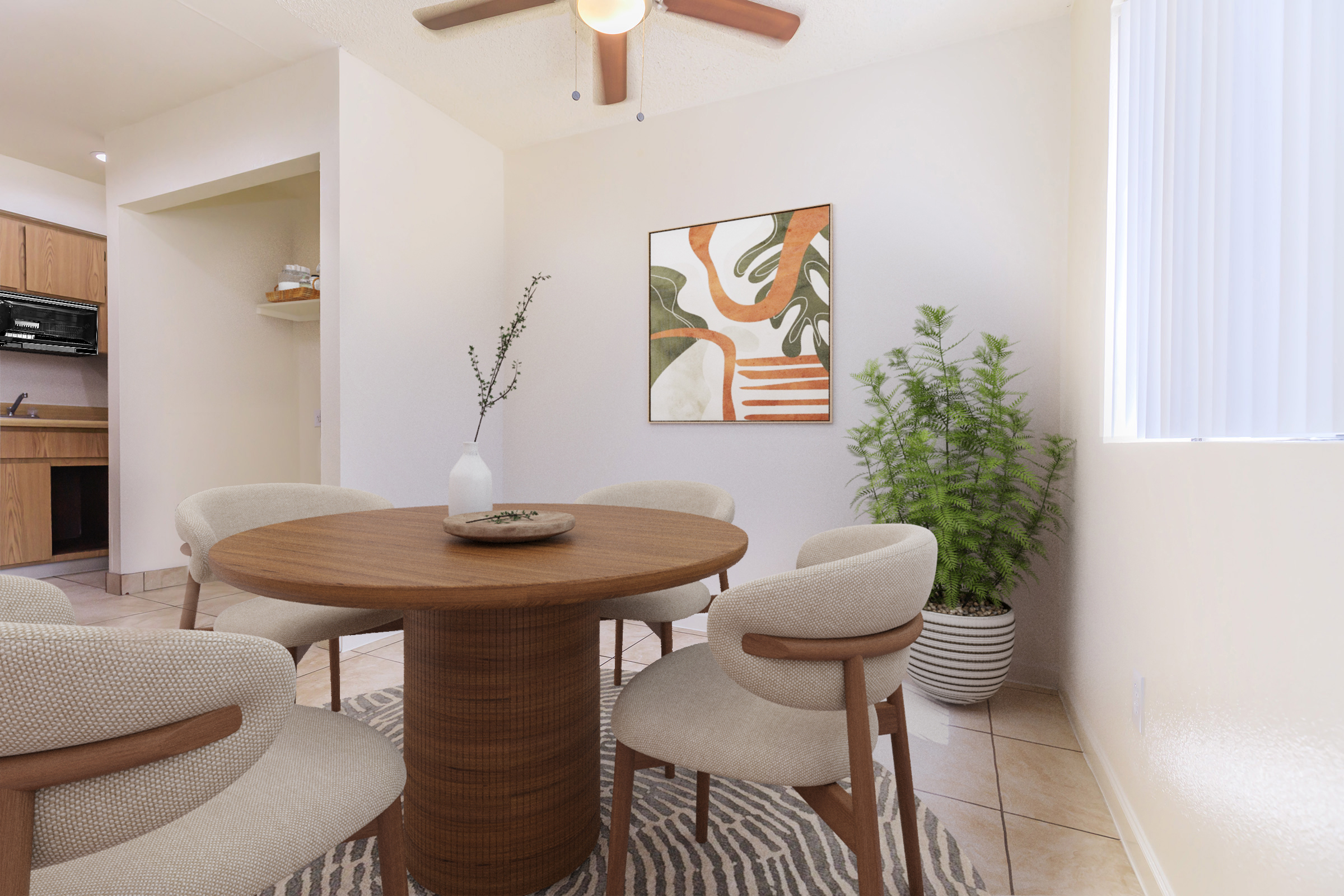 A cozy dining area features a round wooden table surrounded by four beige upholstered chairs. A small vase with branches sits on the table. A potted plant and a colorful wall art piece with abstract shapes and greens can be seen in the background, along with a glimpse of a kitchen area. Natural light filters in through a window.
