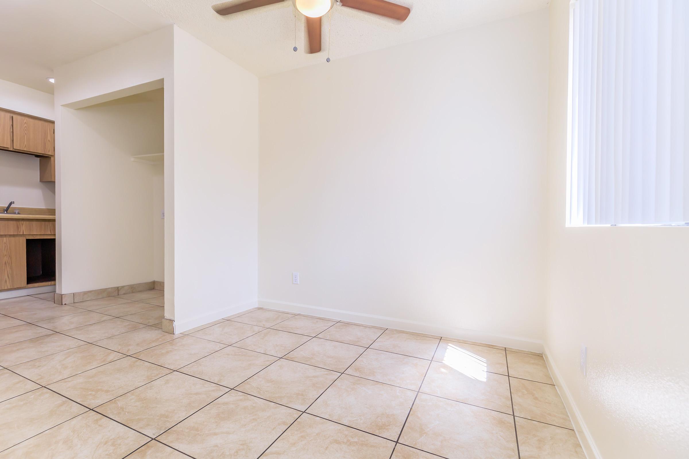 A spacious, empty room featuring tiled floors, light-colored walls, and a ceiling fan. A small archway leads to a kitchen area on the left, and natural light filters in through a window with vertical blinds on the right. The overall atmosphere is bright and airy, ideal for personal customization.