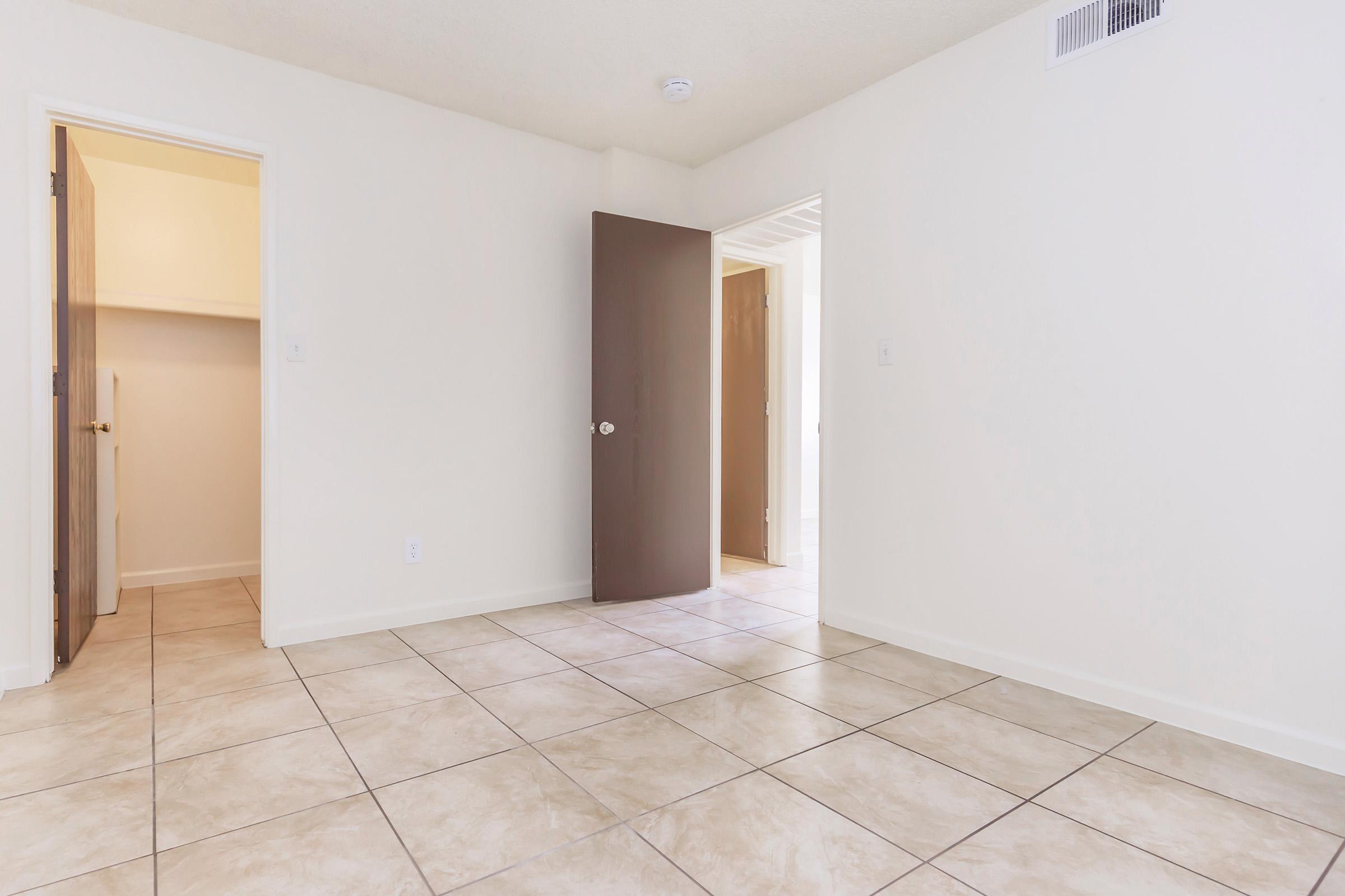 A spacious, empty room with beige tiled flooring. The walls are painted white, and there are two doors: one open leading to a closet and another leading to an adjacent area. Natural light illuminates the space, creating a clean and minimalistic atmosphere.