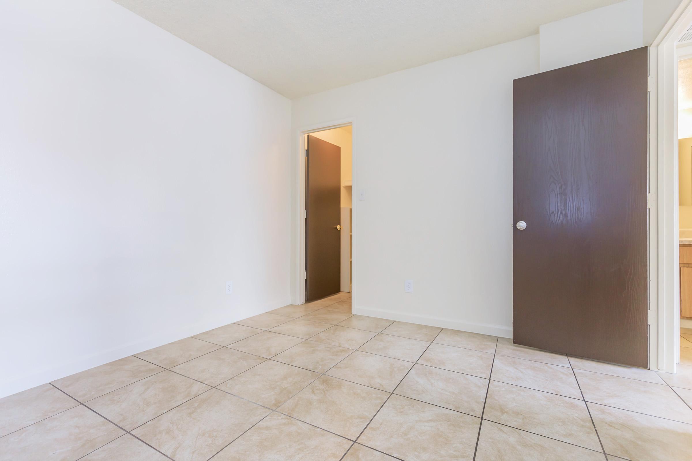 A spacious room with beige tiled floors and plain white walls. The room features a closed dark wood door on the right, leading to another adjacent room, and an open doorway on the left that connects to a bathroom or another area. Natural light fills the space through the doorway.