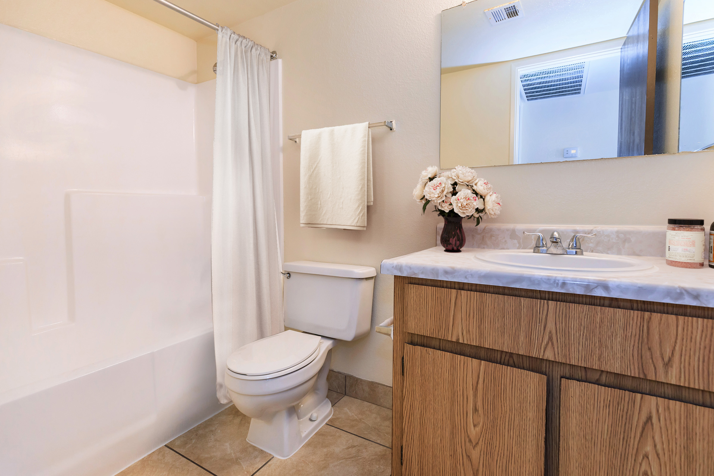 A clean, well-lit bathroom featuring a white bathtub with a shower curtain, a toilet, a wooden vanity with a marble countertop, and a vase of flowers on the sink. The walls are painted in a light color, enhancing the bright atmosphere of the space.
