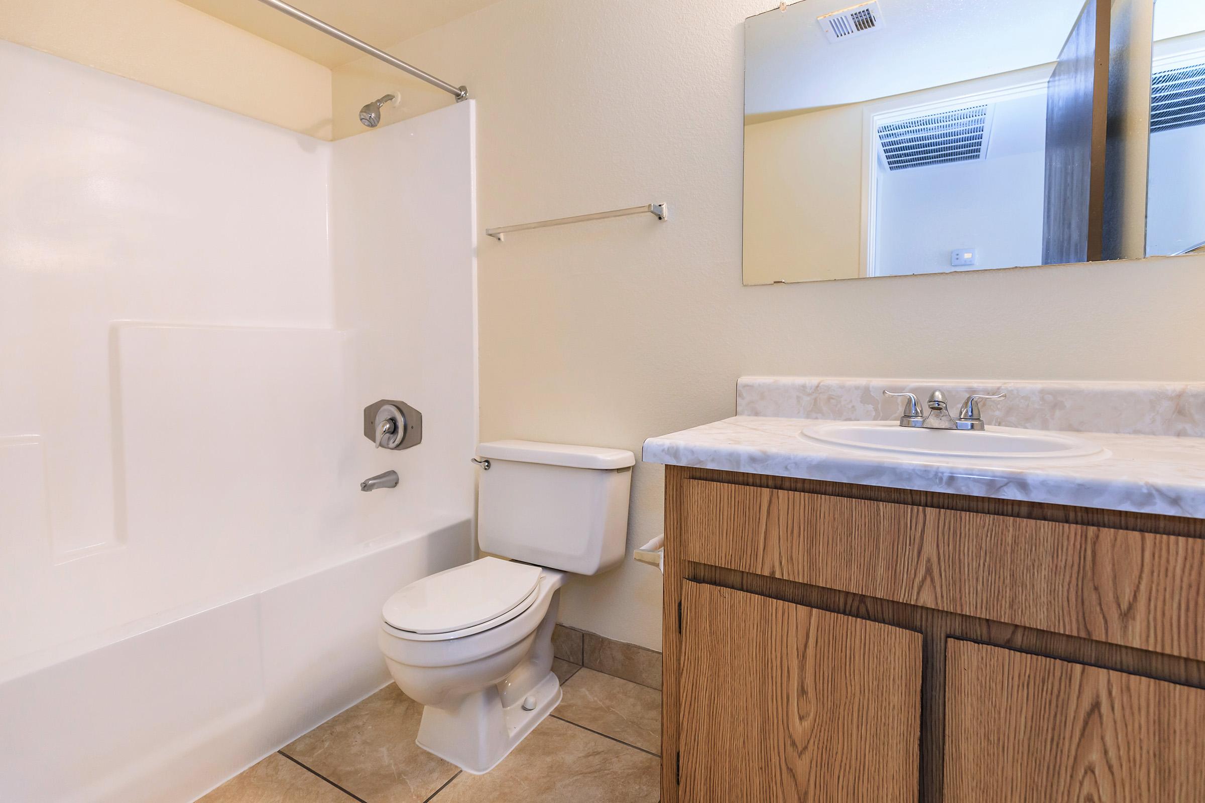 A clean, simple bathroom featuring a white bathtub with a showerhead, a toilet, and a wooden vanity with a marble countertop. A mirror hangs above the sink, and the walls are painted a soft color. Natural light comes from a window above the bathroom fixtures. Tile flooring is visible.