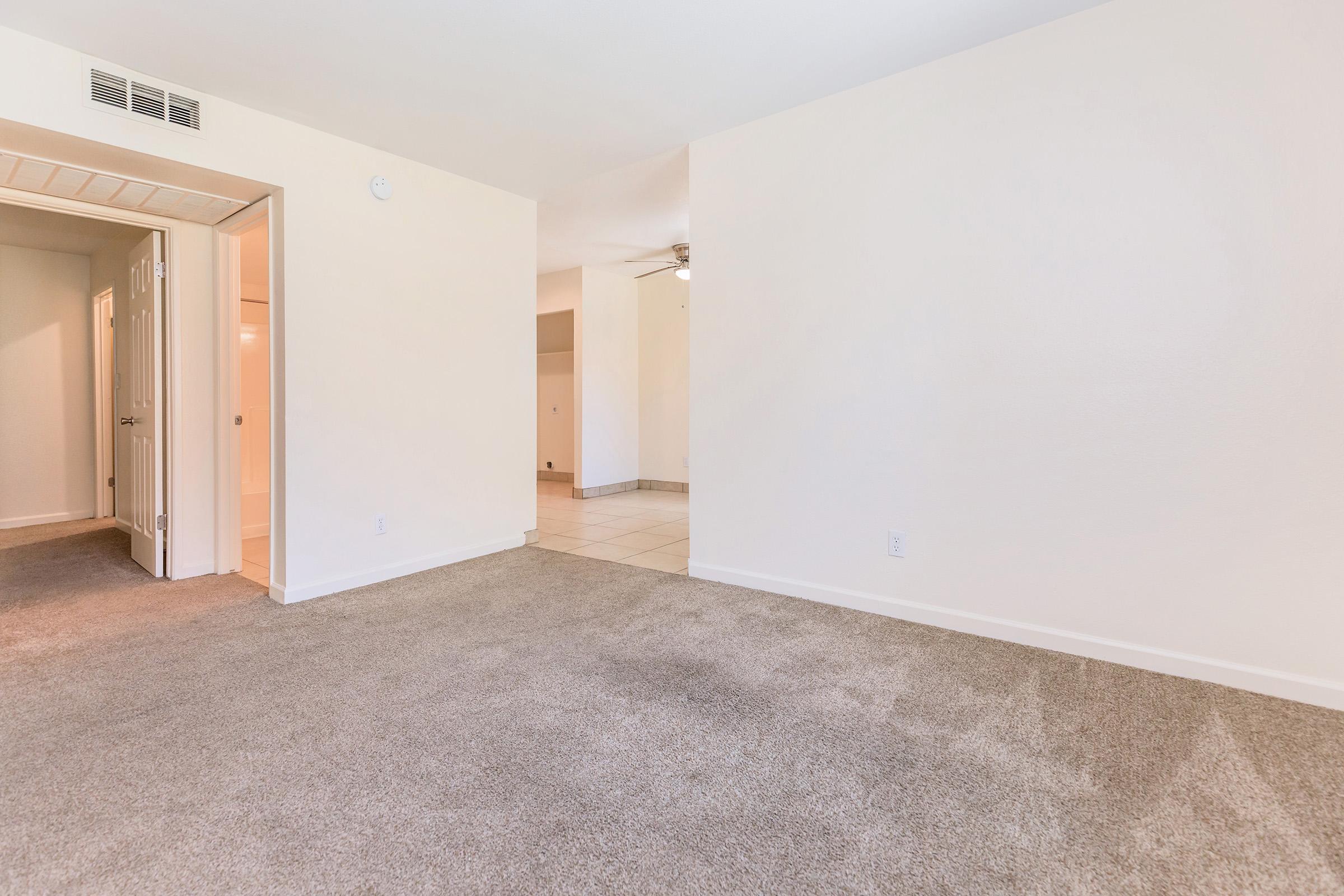 Spacious living area with light-colored walls and beige carpet. A doorway on the left leads to a hallway, while a ceiling fan is visible in the background. The flooring transitions to tile towards the far right. Natural light brightens the room, creating an inviting atmosphere.