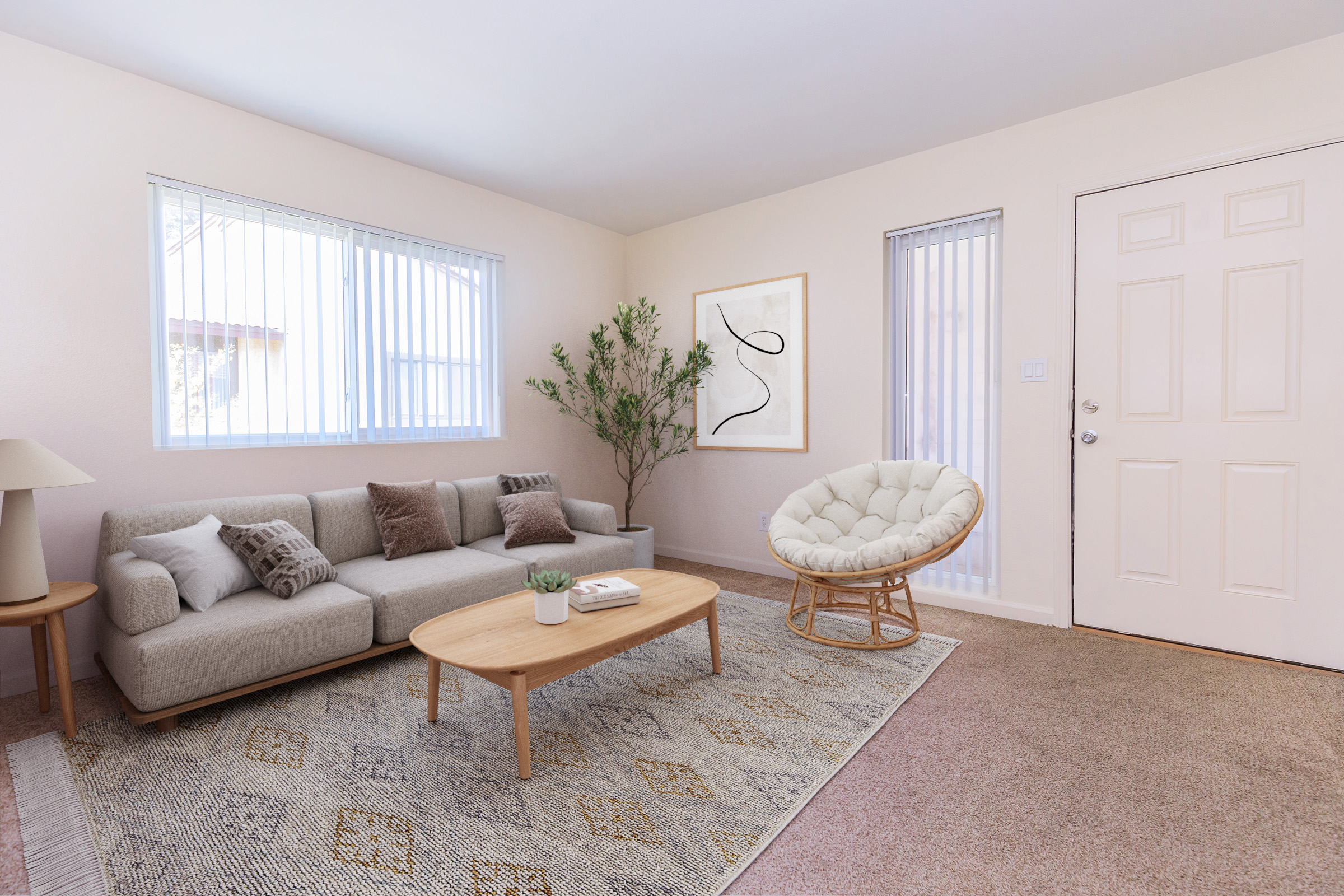 A cozy living room featuring a light-colored sofa with decorative pillows, a round coffee table, and a large, soft area rug. A plant adds a touch of greenery, and there are two windows with blinds. An art piece hangs on the wall, and a door leads outside. The overall atmosphere is bright and inviting.