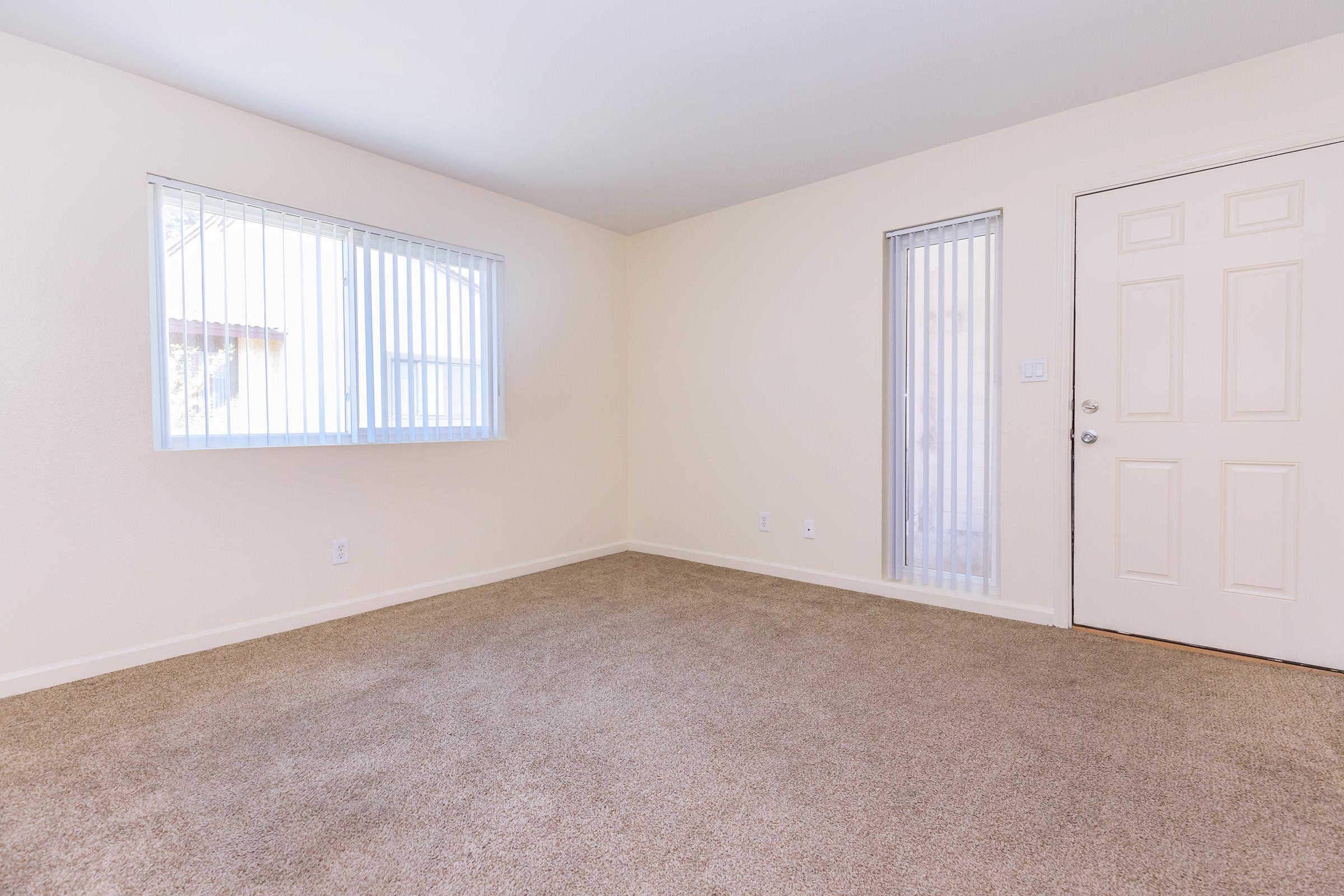 Spacious, empty room with beige walls and light carpet. Two windows with vertical blinds allow natural light, one leading to an entrance door. The overall atmosphere is bright and inviting, ideal for personal decoration or furnishing.