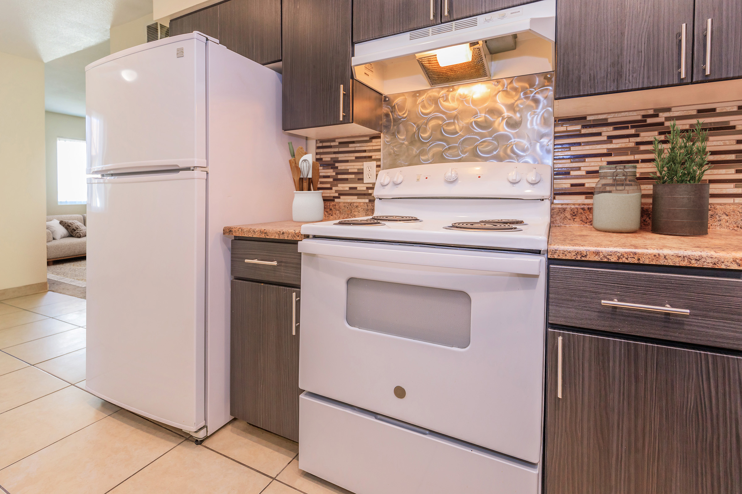 A modern kitchen featuring a white stove, refrigerator, and dark cabinets. The stove has a decorative backsplash with wavy metal design. Countertops are made of speckled granite, and there are potted plants and kitchen utensils on display. The floor is tiled and the area is well-lit.