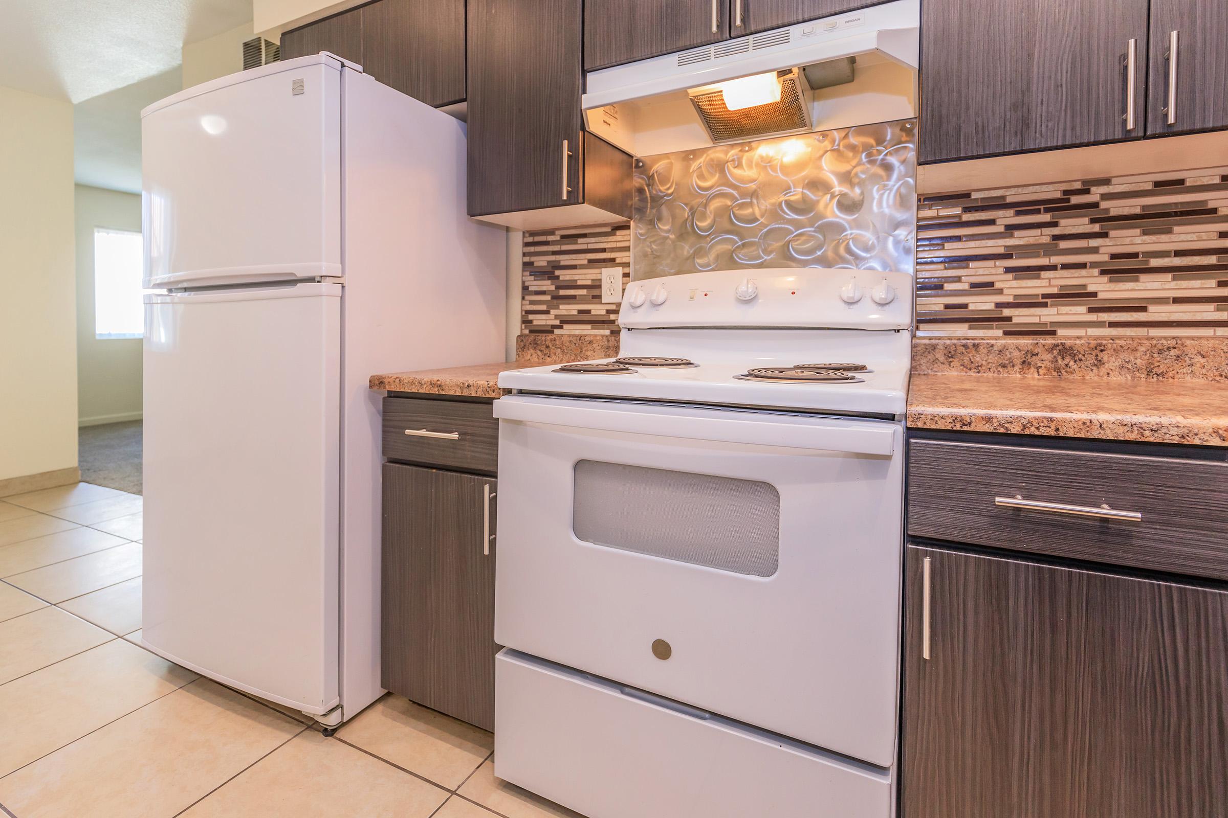 A modern kitchen featuring a white refrigerator, a white electric stove with four burners, and dark wooden cabinets. The backsplash consists of decorative tiles in various shades, and the countertop is made of a light brown stone. Bright lighting enhances the space's clean and inviting look.