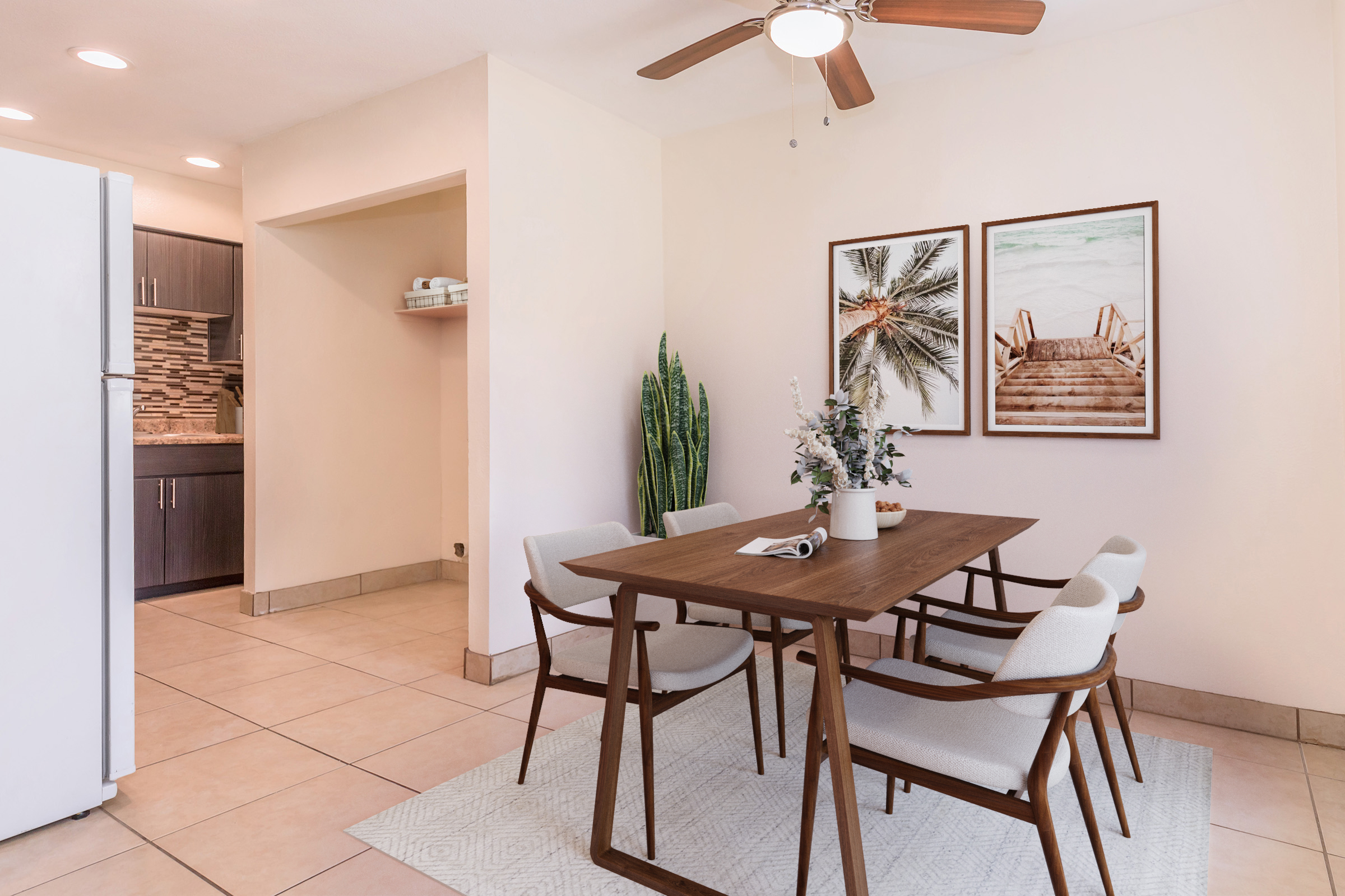 A bright dining area featuring a wooden table with four chairs, a potted plant, and a vase of flowers. The walls are adorned with two framed beach-themed prints. A modern kitchen is visible in the background, along with a white refrigerator and light-colored tiles. The space has a warm, inviting ambiance.