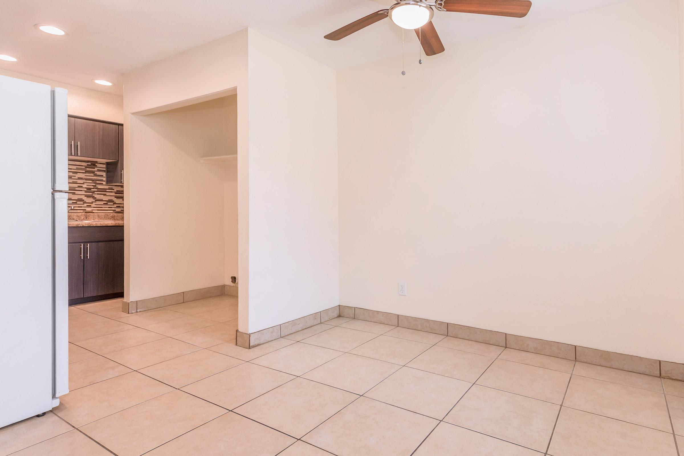 A spacious, unfurnished room with beige walls and tile flooring. A ceiling fan is mounted in the corner, and there is a small opening leading to a kitchen area with dark cabinetry and a decorative tile backsplash. Natural light floods the space from the side.