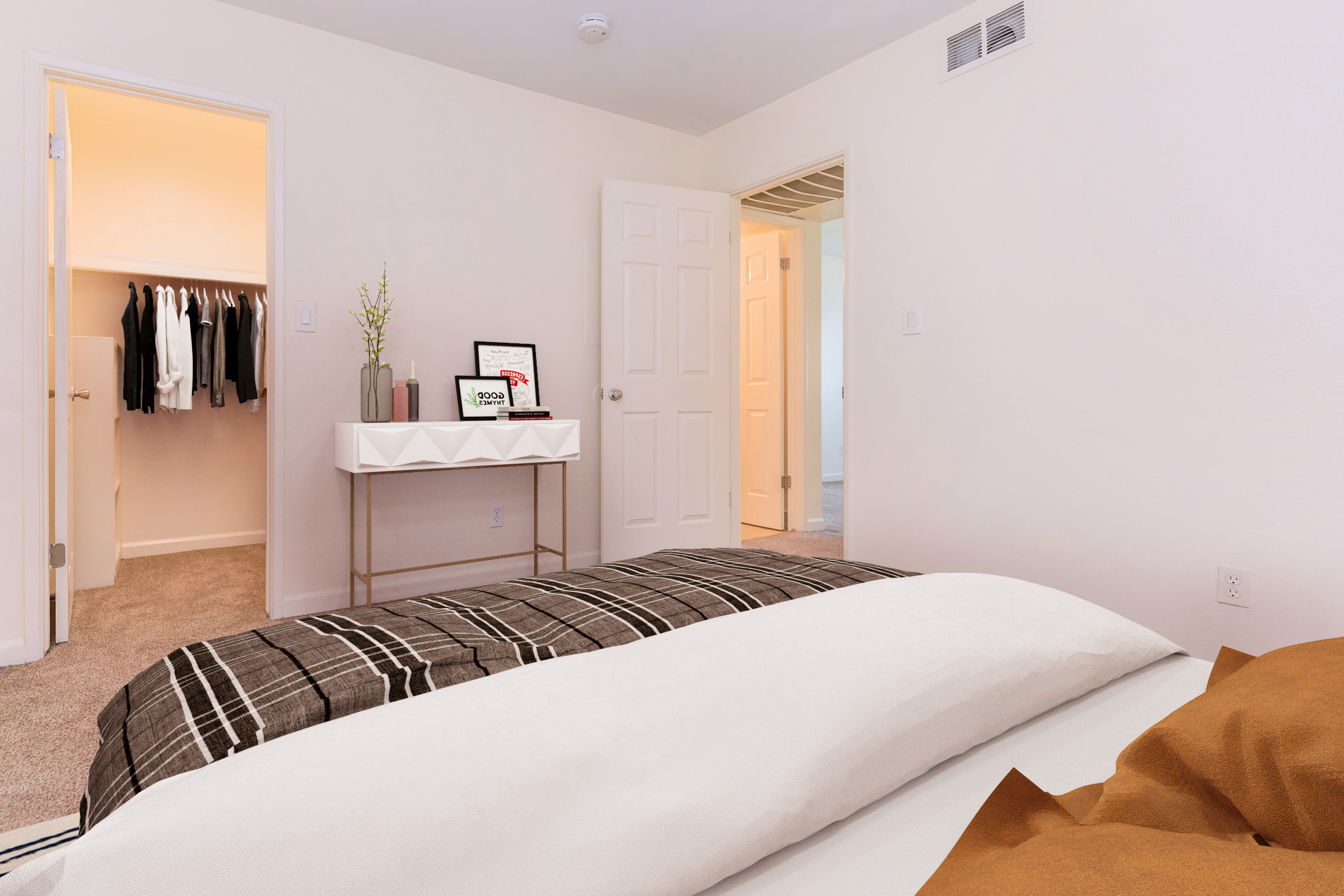 A cozy bedroom featuring a neatly made bed with a checkered blanket. There’s a small desk with a display screen, and an open closet showcasing clothes on hangers. A light-colored wall and soft carpet contribute to a warm ambiance, with a door visible leading to another room.