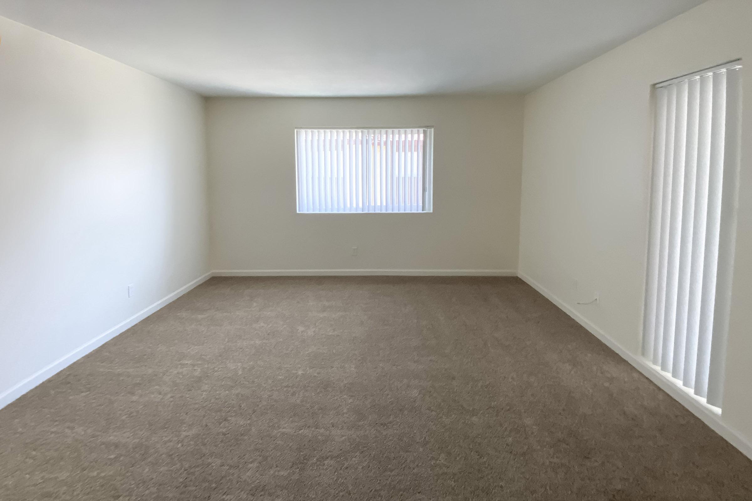 A spacious, empty room with light beige carpet and white walls. There is a window with vertical blinds on one wall, allowing natural light to enter. The room has no furniture, giving it a clean and minimalistic appearance.