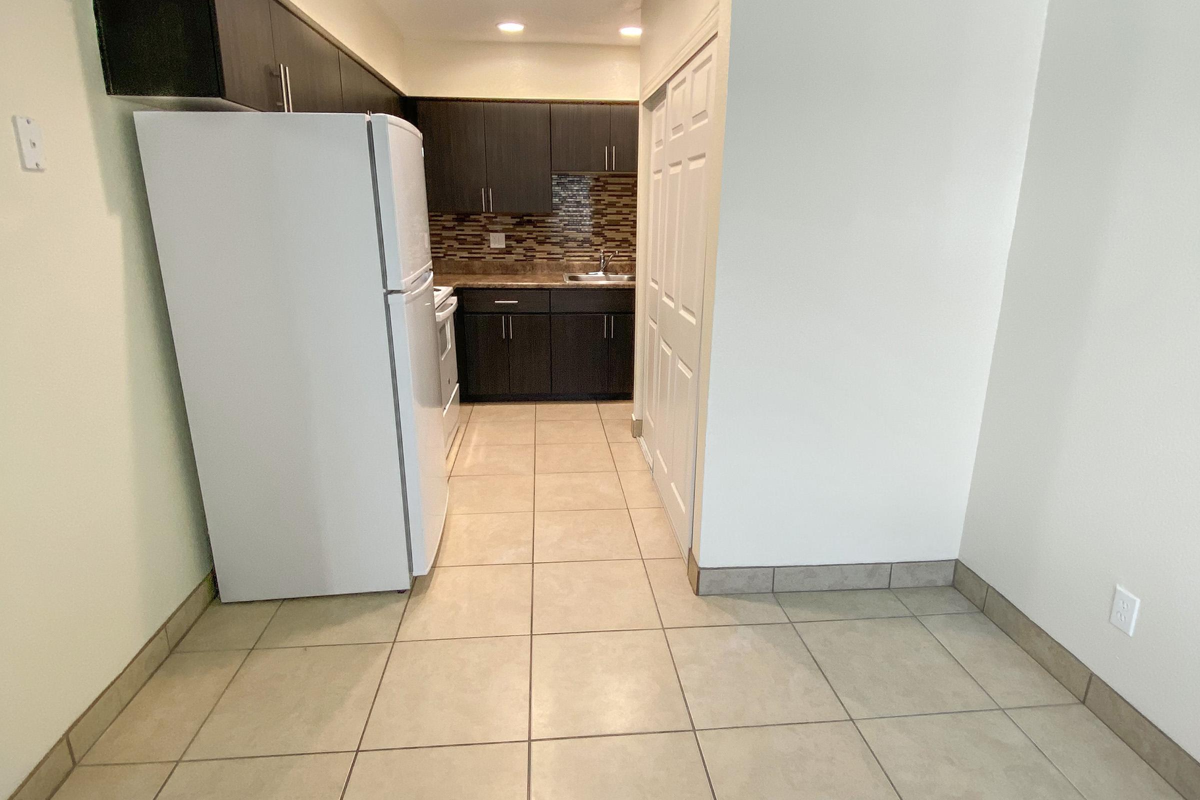 A modern kitchen featuring a white refrigerator and stove, dark wooden cabinets, and a tiled floor. The kitchen has a spacious layout with light-colored walls and a small area leading to a closed door. Decorative backsplash tiles add interest to the design.