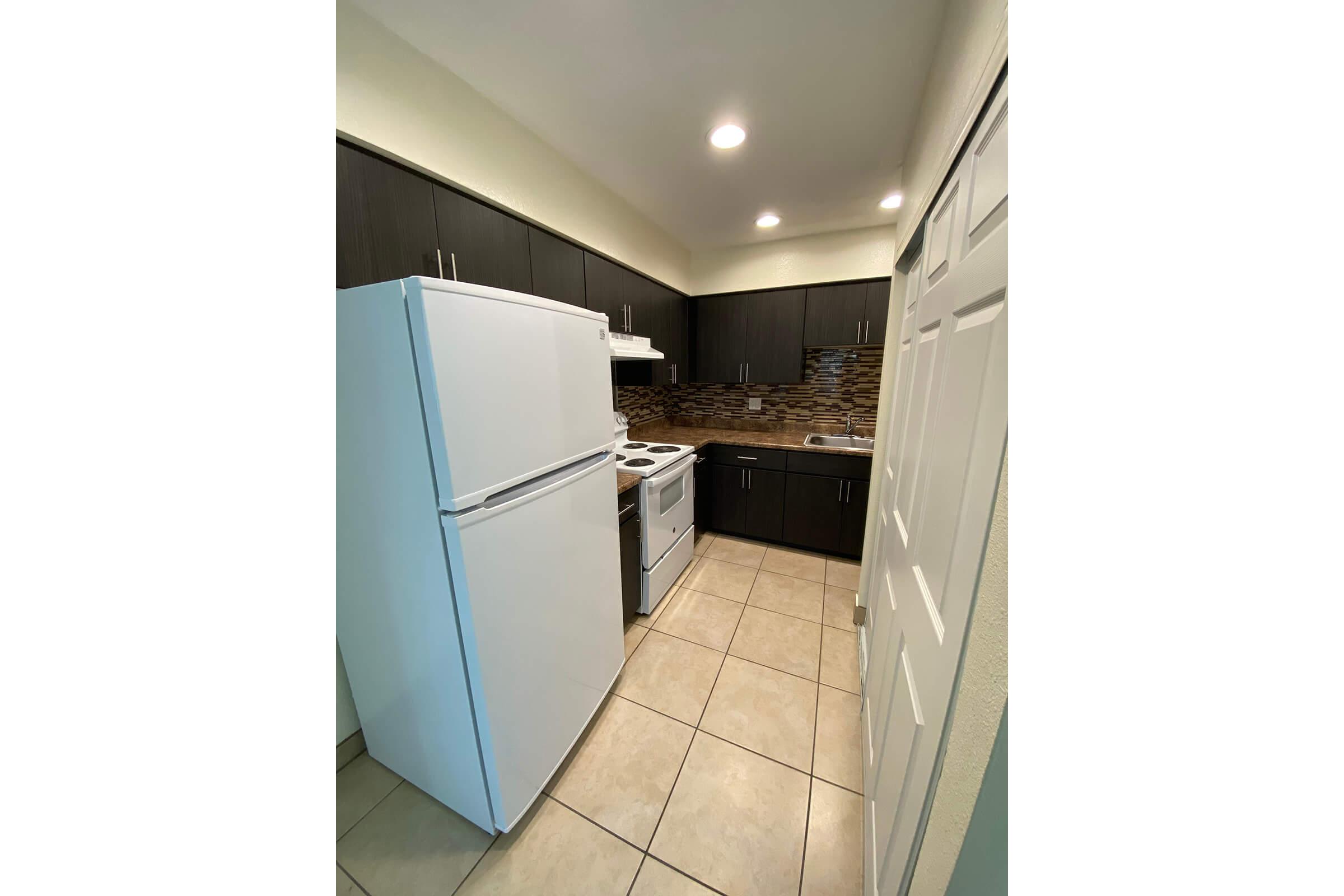 A modern kitchen featuring dark wooden cabinets, a white refrigerator, and a white stove with an oven. The backsplash consists of decorative tiles in earthy tones, and the floor is tiled in light color. The kitchen includes a small amount of countertop space and overhead cabinets for storage.