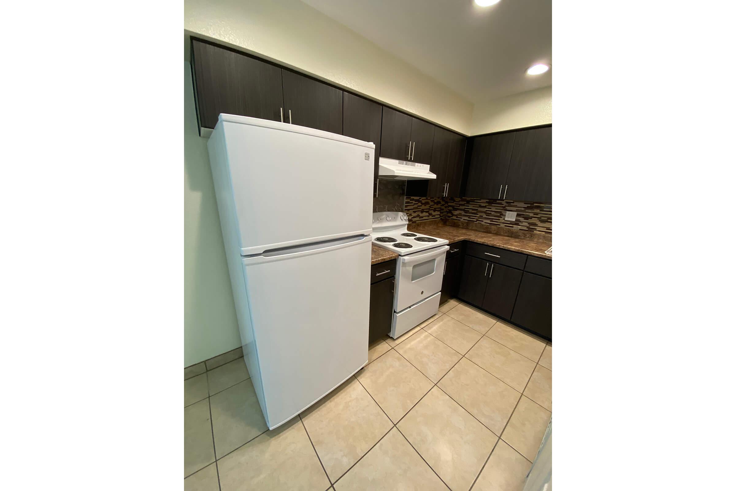 A modern kitchen featuring dark wood cabinets, a white refrigerator, a white stove, and a white oven. The countertops are complemented by a tiled backsplash in neutral tones. The flooring consists of light beige tiles, creating a clean and spacious environment. Bright lighting enhances the overall ambiance.