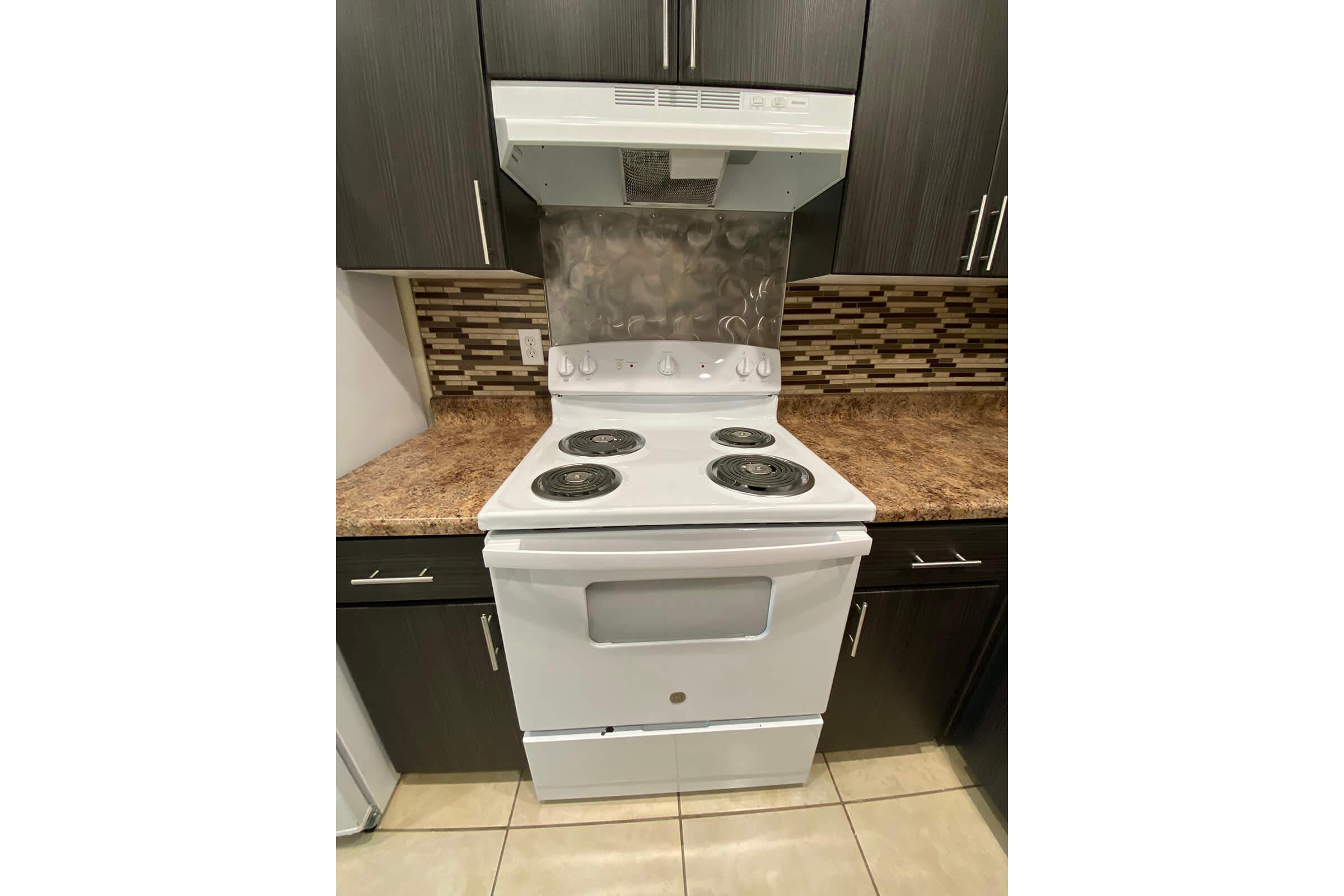 A white kitchen stove with four burners and an oven, situated in a modern kitchen. The stove has a stainless steel hood above it and is placed against a backsplash made of rectangular tiles in various shades. The countertop is brown and the cabinets are dark wood.