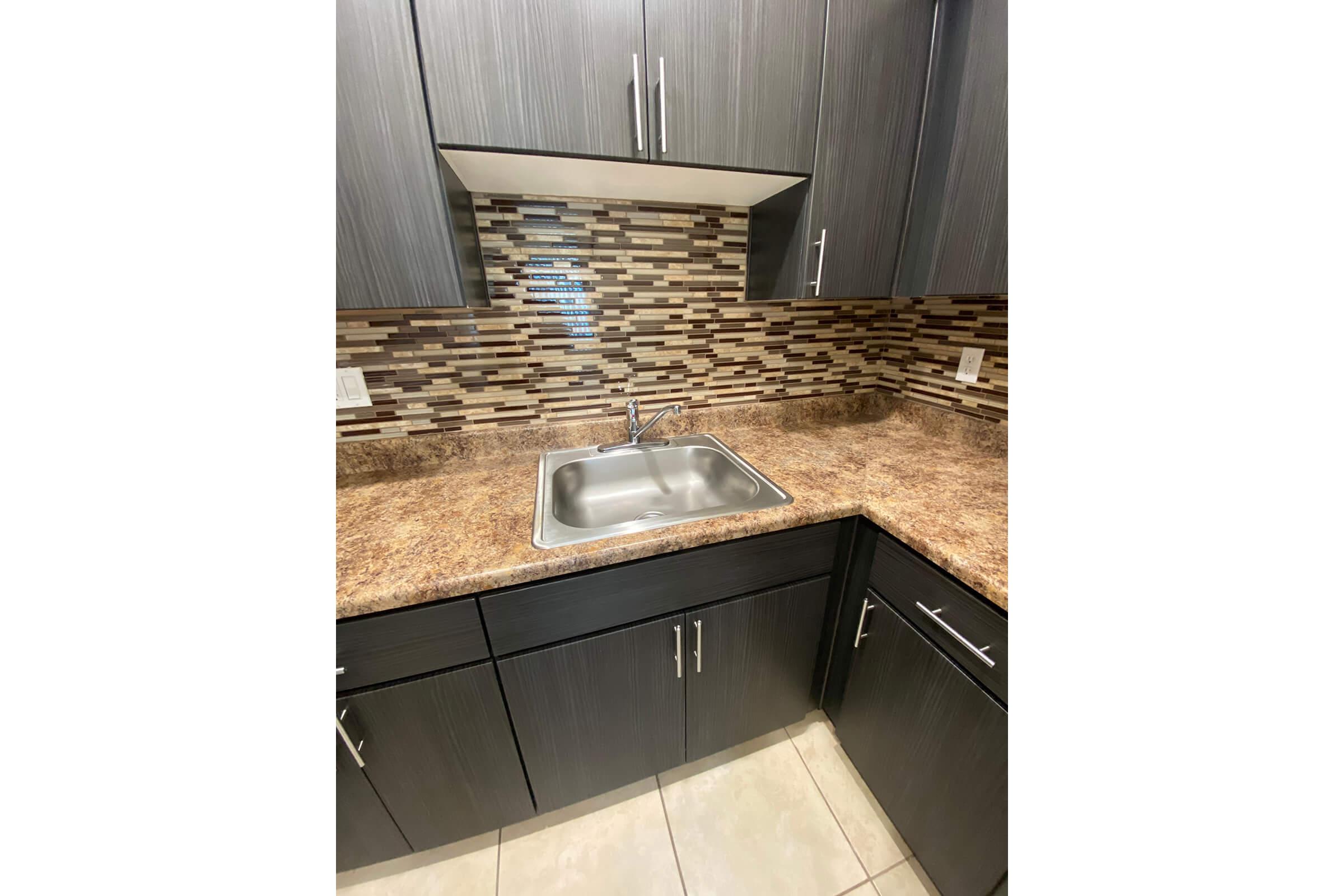 A modern kitchen space featuring dark cabinetry and a stylish mosaic backsplash. The countertop is made of a light-colored material with a stainless steel sink. The floor has large tiles in a neutral shade, creating a clean and contemporary look.